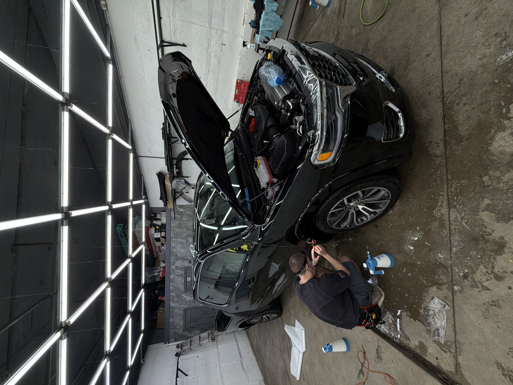 The image captures an interior view of an automotive detailing and film application facility in Krasnoyarsk, Russia. A large, black, four-door RAM 1500 pickup truck is the central subject, with its hood fully open, revealing the engine compartment. A clear plastic water bottle rests on a component within the engine bay.
An adult male, wearing a dark t-shirt, dark shorts with a patch resembling the Russian flag, a headlamp, and safety glasses, kneels on the wet, light grey concrete floor next to the front passenger side wheel arch of the truck. He is actively engaged in applying or trimming a clear film, using a small orange squeegee-like tool and a utility knife. The floor around him is visibly wet, indicating a fluid-assisted application process. Two blue-and-white pump spray bottles are positioned on the floor near the worker. A partially unrolled roll of white film material and several discarded clear film pieces are also on the floor.
The facility is brightly illuminated by an overhead grid system of linear fluorescent or LED lights on a dark ceiling. The walls are constructed of white, textured concrete blocks, with a section of dark grey cladding on the rear wall. High shelving units above this cladding are cluttered with various boxes, including one labeled "SF film" and "DMKPRO", and miscellaneous automotive parts. Black metal shelving, laden with automotive components, is mounted on the right wall, adjacent to a table holding detailing supplies and blue cloths. An aluminum extension ladder leans against the left wall. A visible drainage channel runs along the concrete floor in the foreground.
