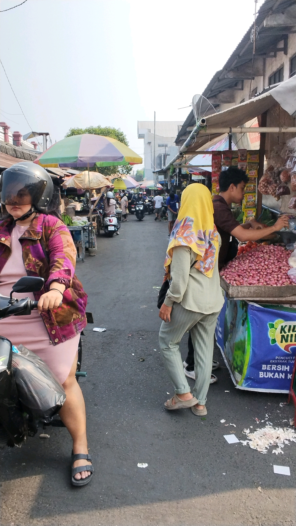 This is a medium-shot, eye-level photograph taken outdoors in the city of Mojokerto, Indonesia, likely in a marketplace. The scene depicts a busy street with stalls and pedestrians. In the foreground on the left, a woman wearing a pink dress and a patterned jacket is sitting on a motorcycle, her face obscured by a dark helmet. Her left leg, clad in a pink dress, is extended, and she wears dark sandals. A large black plastic bag is attached to the front of the motorcycle. To the right of the motorcycle, a person wearing a yellow hijab and a light green outfit with striped pants walks away from the camera. This person is wearing tan sandals. Behind them, a man with dark hair, wearing a brown shirt, is standing behind a market stall laden with red onions. The stall has a blue banner with green accents that features Indonesian text. In the background, more market stalls with umbrellas and awnings are visible, along with several motorcycles and people moving around. The ground is paved with asphalt, and there is some litter scattered on it. The sky is bright and clear.