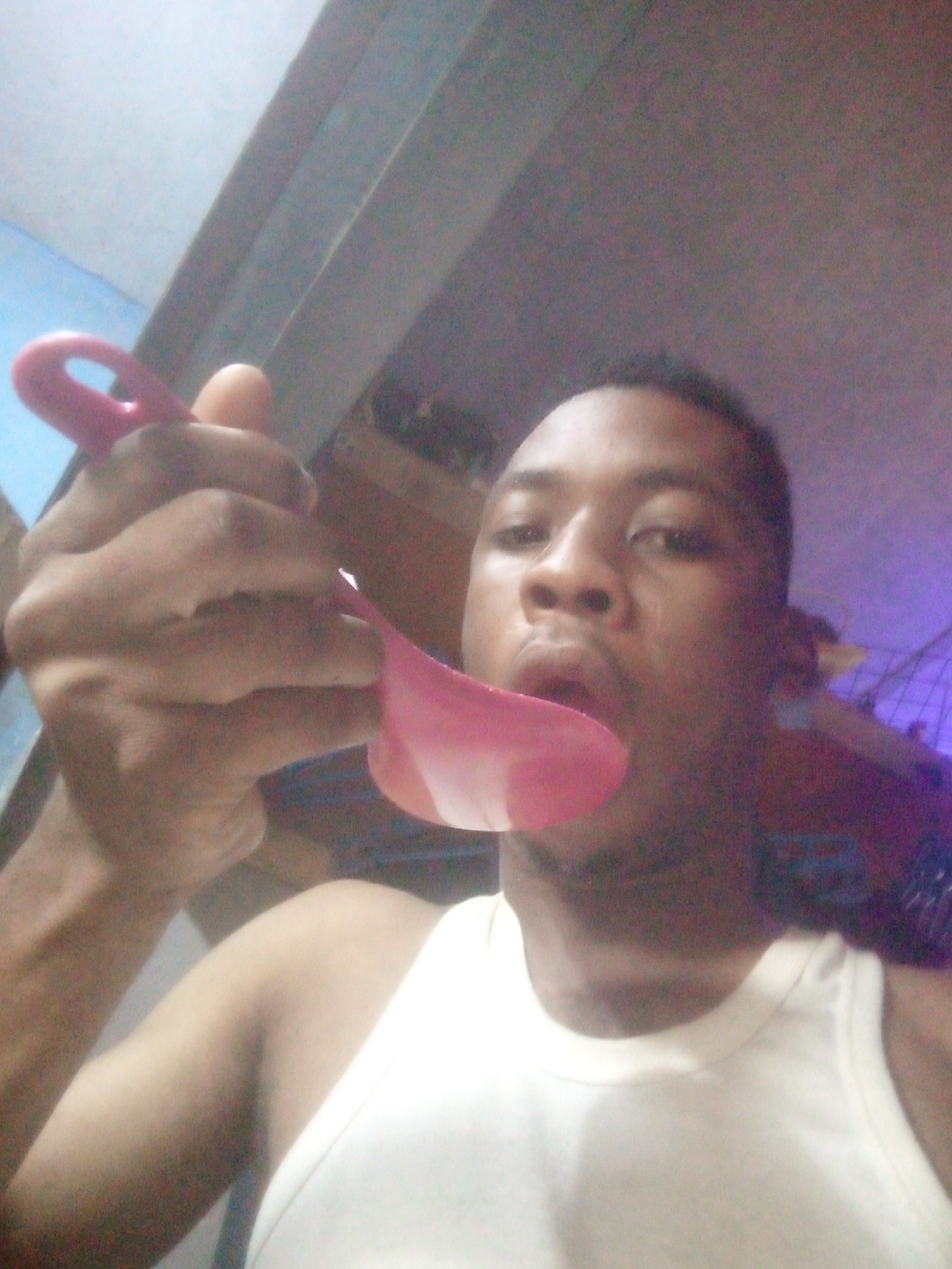 A man with short dark hair and dark skin is holding a pink spoon to his mouth. He is wearing a white tank top. The background appears to be an indoor space, possibly a home, with a blue wall visible in the upper left. The image quality is somewhat grainy.