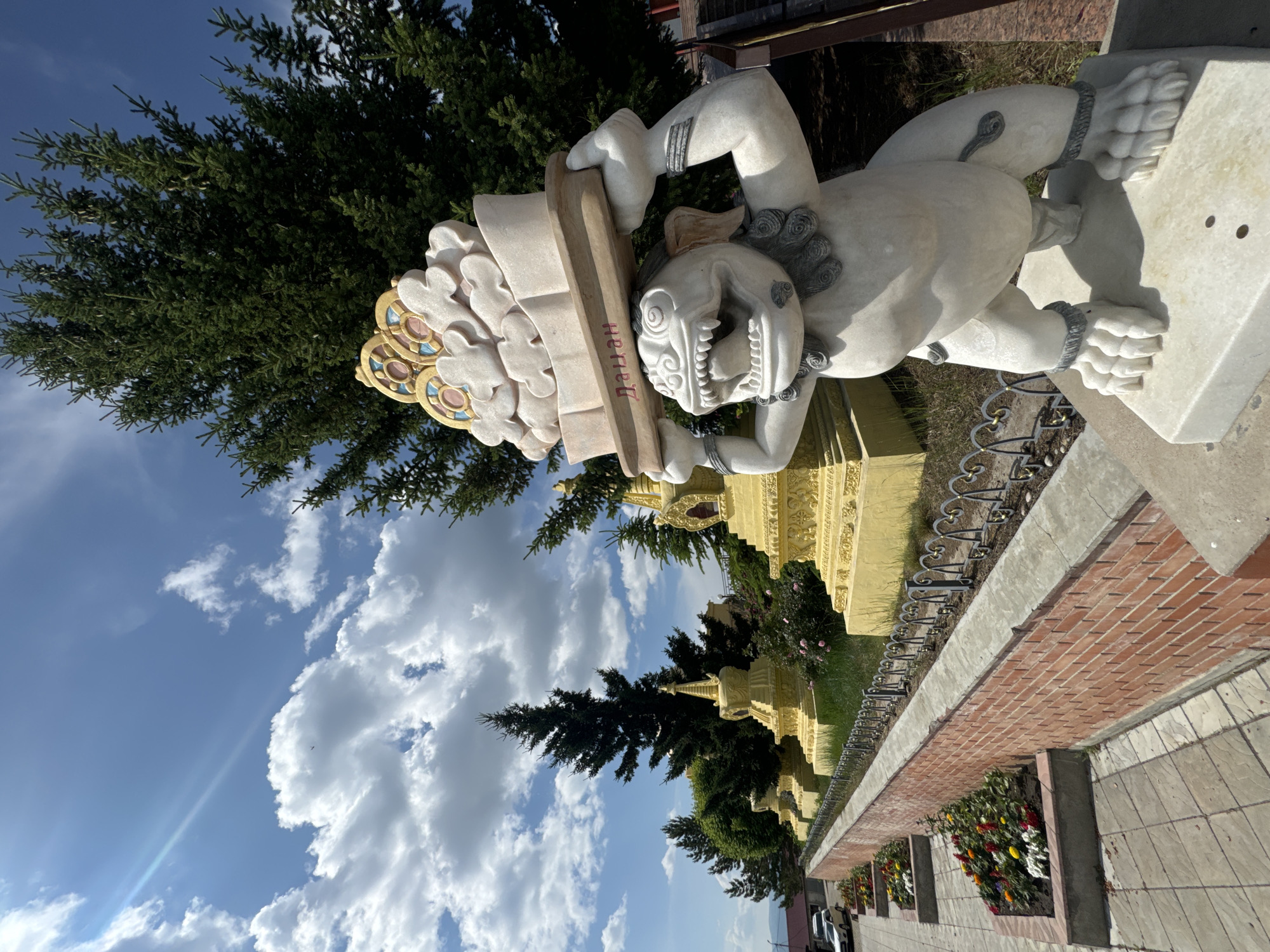 The image shows a statue of a mythical creature carrying a decorative offering on its head. The creature is white, with sharp teeth, claws, and adorned with necklaces and bracelets. The offering appears to be made of stone, with a wooden base and colorful decorations on top. The statue is positioned in front of a row of golden stupas, which are Buddhist religious monuments. The stupas are of varying sizes and are surrounded by greenery, including tall evergreen trees. The foreground features a brick wall and a paved walkway with flowerbeds. In the background, there are some buildings and a blue sky with white clouds. The overall scene suggests a religious or cultural site. The statue is identified as "Дацан", which is a name for a Buddhist monastery in Buryatia, Russia. Therefore, the location is a Buddhist monastery in Ulan-Ude, Russia.