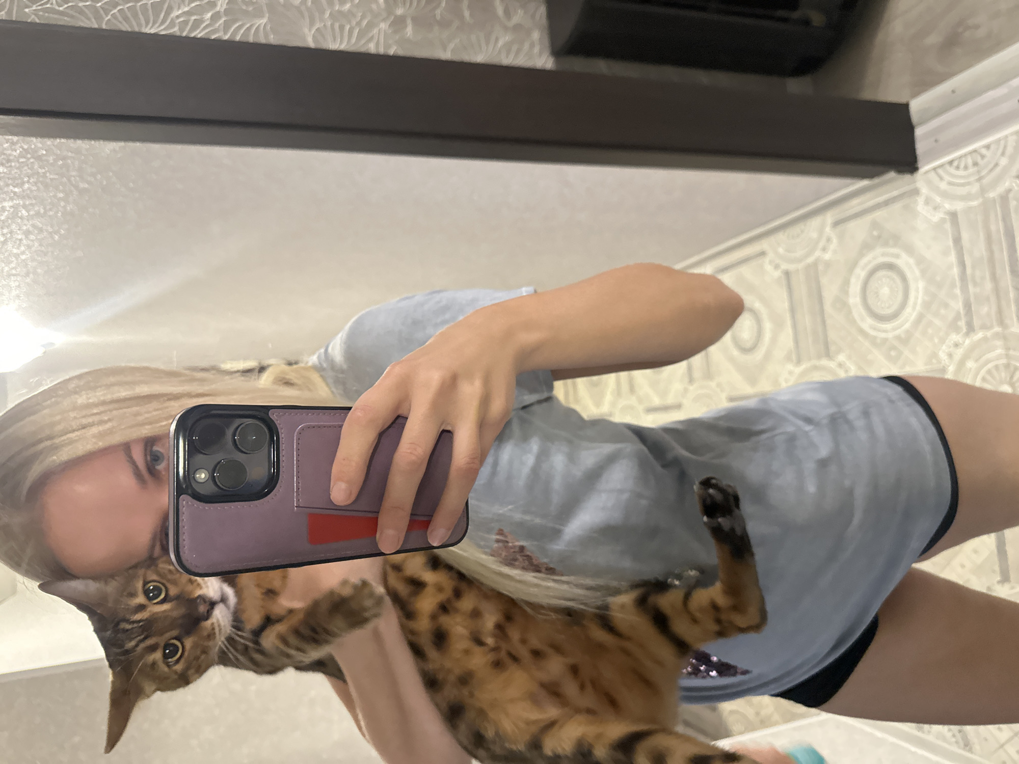 A woman with blonde hair holds a Bengal cat while taking a selfie in a room with patterned wallpaper and a tiled floor. The woman is wearing a light blue t-shirt and black shorts. The cat is brown with black spots. The environment appears to be indoors. The image does not provide any specific information about the city of Energetik, Russia.