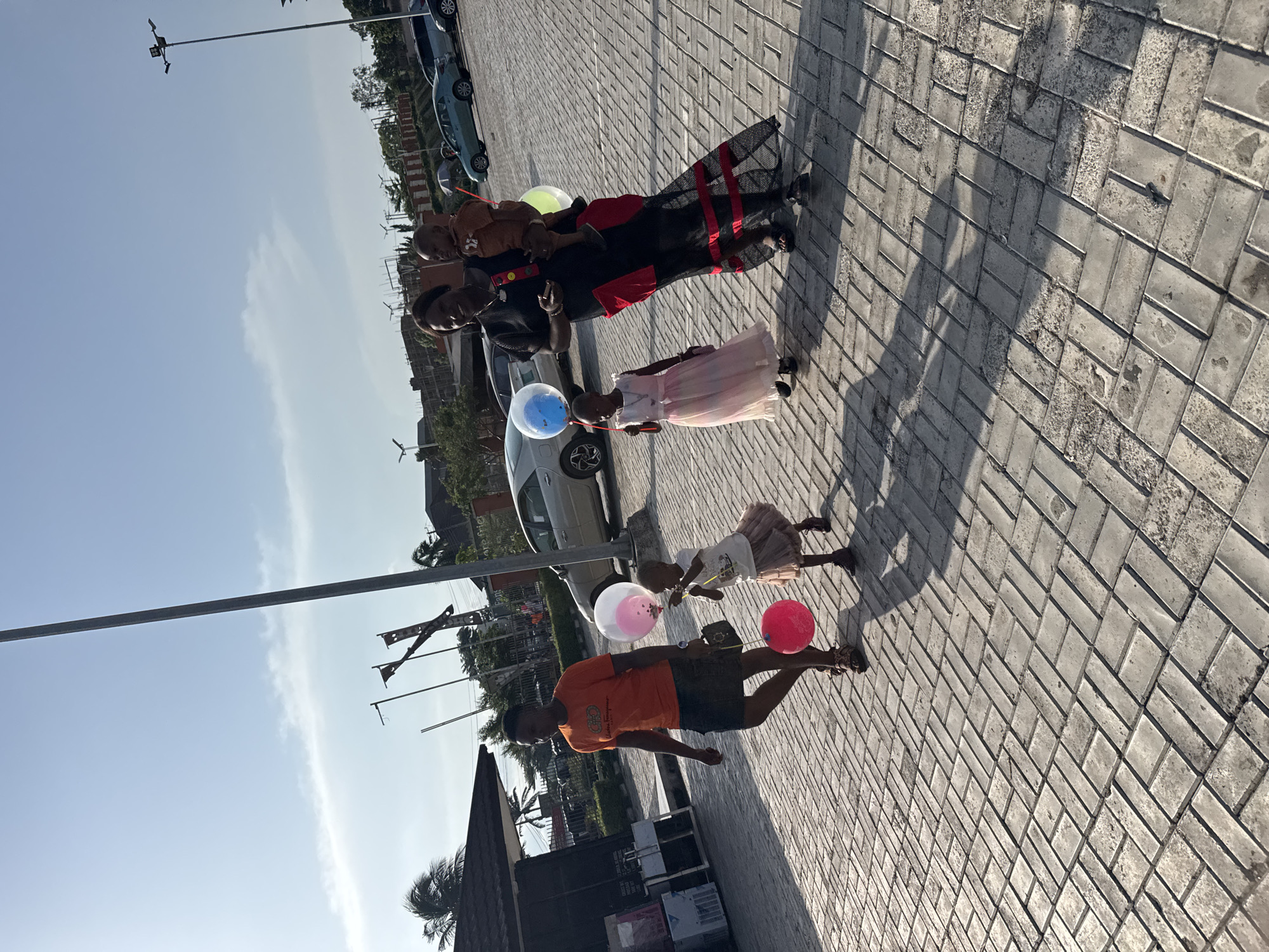 The image depicts a family standing on a brick-paved area in Onitsha, Nigeria. There are four individuals: an adult woman in a black and red dress holding a child, a young girl in a pink dress, and a younger child with another adult. Each person holds a balloon. The environment includes cars parked in the background, light poles, and some greenery, suggesting an urban or suburban setting. The sky is clear. The overall atmosphere seems casual.