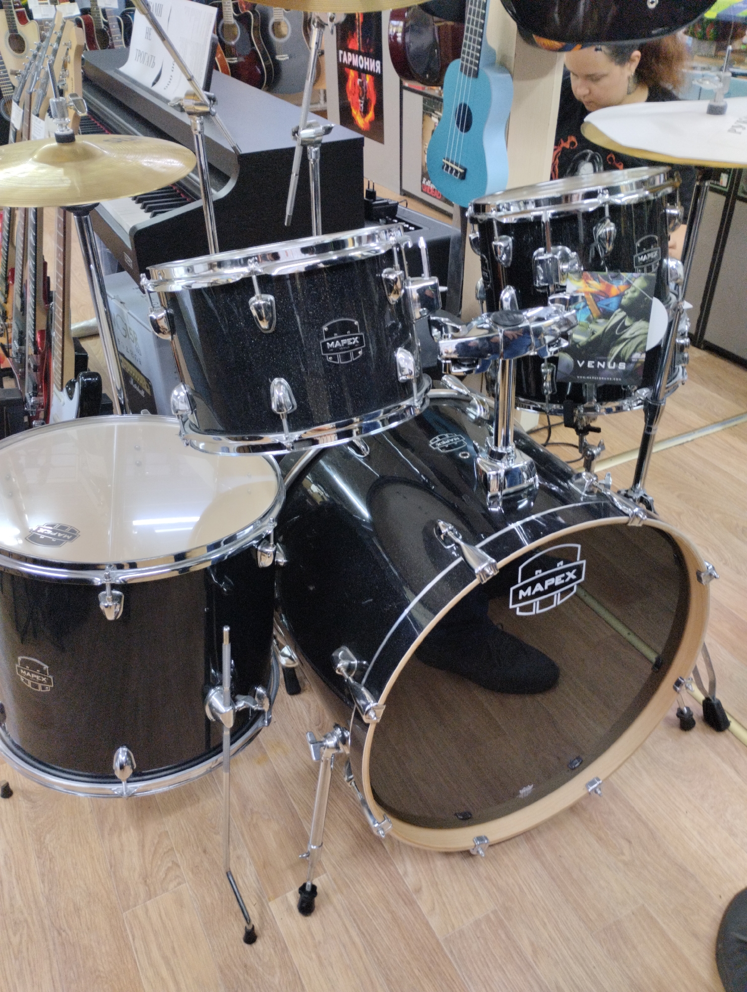 An indoor medium shot captures a Mapex drum set with a black sparkle finish in a retail environment. The set includes a bass drum, a snare drum, and likely other toms though they are partially obscured. The drums are equipped with chrome hardware. A cymbal is visible above the snare drum, suspended by a stand. The floor is made of light-colored wood planks. In the background, a person with dark, curly hair is looking down, partially visible behind the drum set. To the left of the drums, a black keyboard is positioned. Various musical instruments, including guitars and a blue ukulele, are displayed on racks in the background. Posters with text and graphics are also visible on the walls. The overall scene suggests a music store in Kostanay, Kazakhstan.