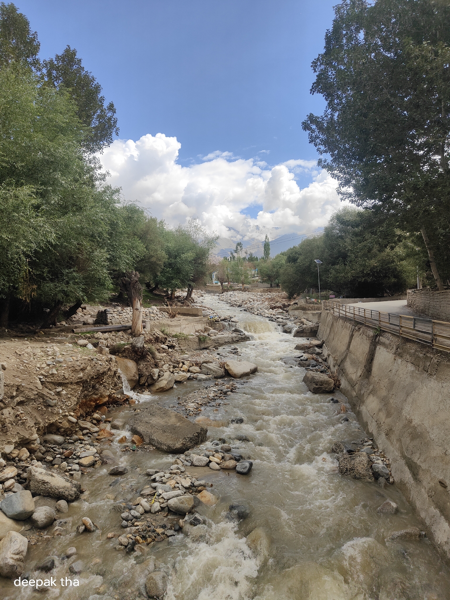 A wide-angle, eye-level shot captures a turbulent, muddy river flowing through a rocky streambed. Dense green trees line the left bank, their foliage casting shadows on the uneven, stony ground. On the right bank, a concrete embankment with a wooden railing separates the river from a paved road. The sky above is a vibrant blue, dotted with fluffy white clouds. In the background, buildings and more trees are visible, with mountains in the distance. The scene suggests a natural landscape with some human infrastructure. The watermark "deepak tha" is visible in the lower left corner.