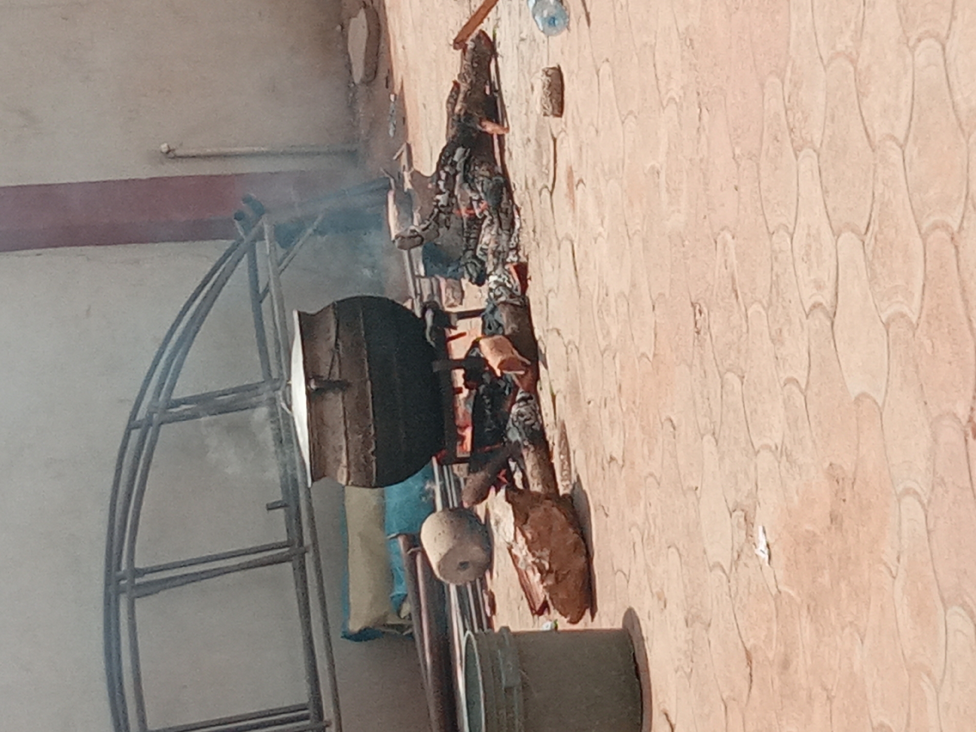 The image captures an outdoor cooking scene, likely in a courtyard or open space in Sabon Tasha, Nigeria, under bright daylight. The central focus is a large, dark-colored metal pot or cauldron, which is placed over an active open fire. Smoke visibly rises from the burning wood and charcoal, indicating that cooking or heating is underway.
Scattered on the ground around the fire are various objects: a spherical, light-colored item, possibly a gourd or a large stone, and a substantial, irregularly shaped brown object, which could be wood or an item being prepared. Further to the right, a discarded plastic water bottle with a blue cap and a rectangular, brick-like object are visible. A blue and white striped bag or piece of fabric is partially seen behind the pot.
The ground is paved with reddish-brown tiles, featuring a distinctive scalloped or scale-like pattern. To the left of the cooking setup, a tall, curved metal frame structure, resembling scaffolding or a support frame, stands against a light-colored wall. In the foreground on the bottom right, a dark grey cylindrical object, possibly a bucket or a section of a pipe, is present. The background wall on the left is plain and light-colored, with a horizontal reddish-brown beam and a white pipe attached to it. The bright illumination suggests a sunny daytime setting. No individuals or visible text are depicted in the image.