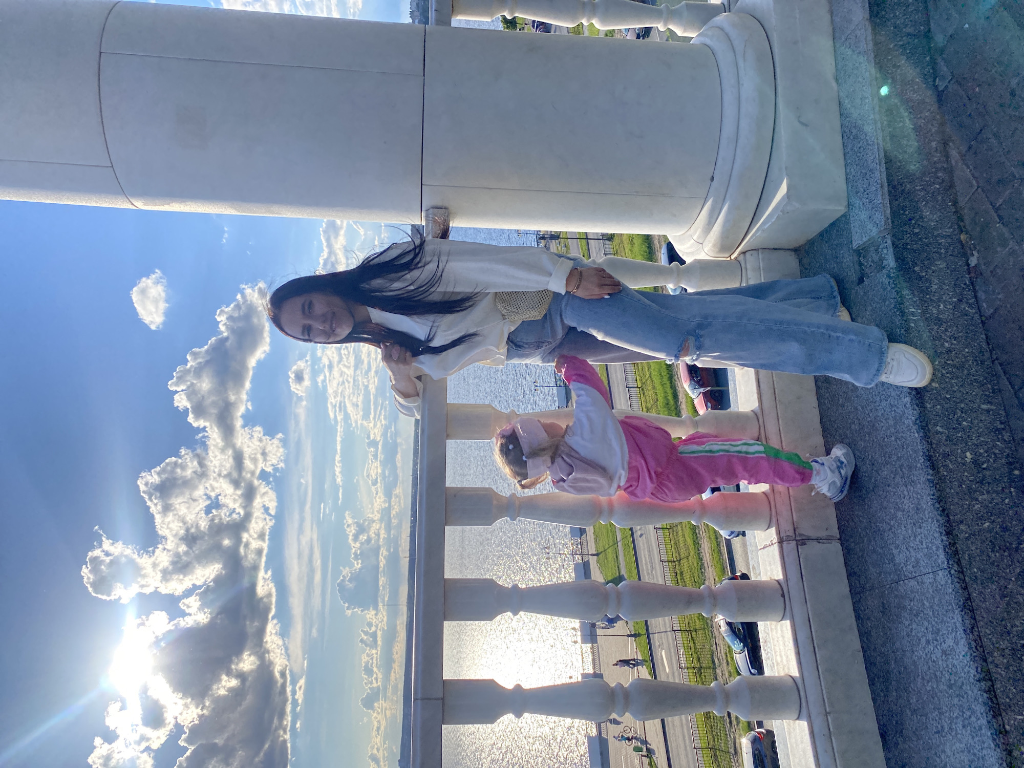 This image, when viewed upright, portrays an adult woman and a young girl on a scenic promenade, likely overlooking the Izhevsk Pond in Izhevsk, Russia. The scene is illuminated by the late afternoon or early evening sun, creating sparkling reflections on the water and a noticeable lens flare.
In the foreground, a white, classical stone balustrade with several ornate pillars frames the view. The ground beneath the subjects is a dark, speckled grey pavement. A young woman with long, dark hair stands smiling at the camera. She is dressed in a loose-fitting, light-colored long-sleeved shirt, light blue ripped jeans, and white sneakers. Her left hand is gently holding the hand of a young girl standing next to her.
The child, positioned to the adult's right, wears a bright pink tracksuit with green stripes along the sides, and light-colored sneakers with blue accents. She appears to have light-colored hair and possibly a hat or head covering. Her gaze is directed away from the camera.
Beyond the balustrade, a large body of water, likely a lake or wide river, shimmers with sunlight. In the distance, a road or pathway runs along the water's edge, with a few vehicles and indistinct figures visible. The sky is a clear blue, dotted with scattered white, cumulus clouds. The low sun is partially obscured by clouds, casting a strong glow and creating a visible lens flare effect in the lower left portion of the sky.
The image conveys a sense of happiness and leisure, capturing a pleasant outdoor moment between the two individuals. There is no visible text in the media file.