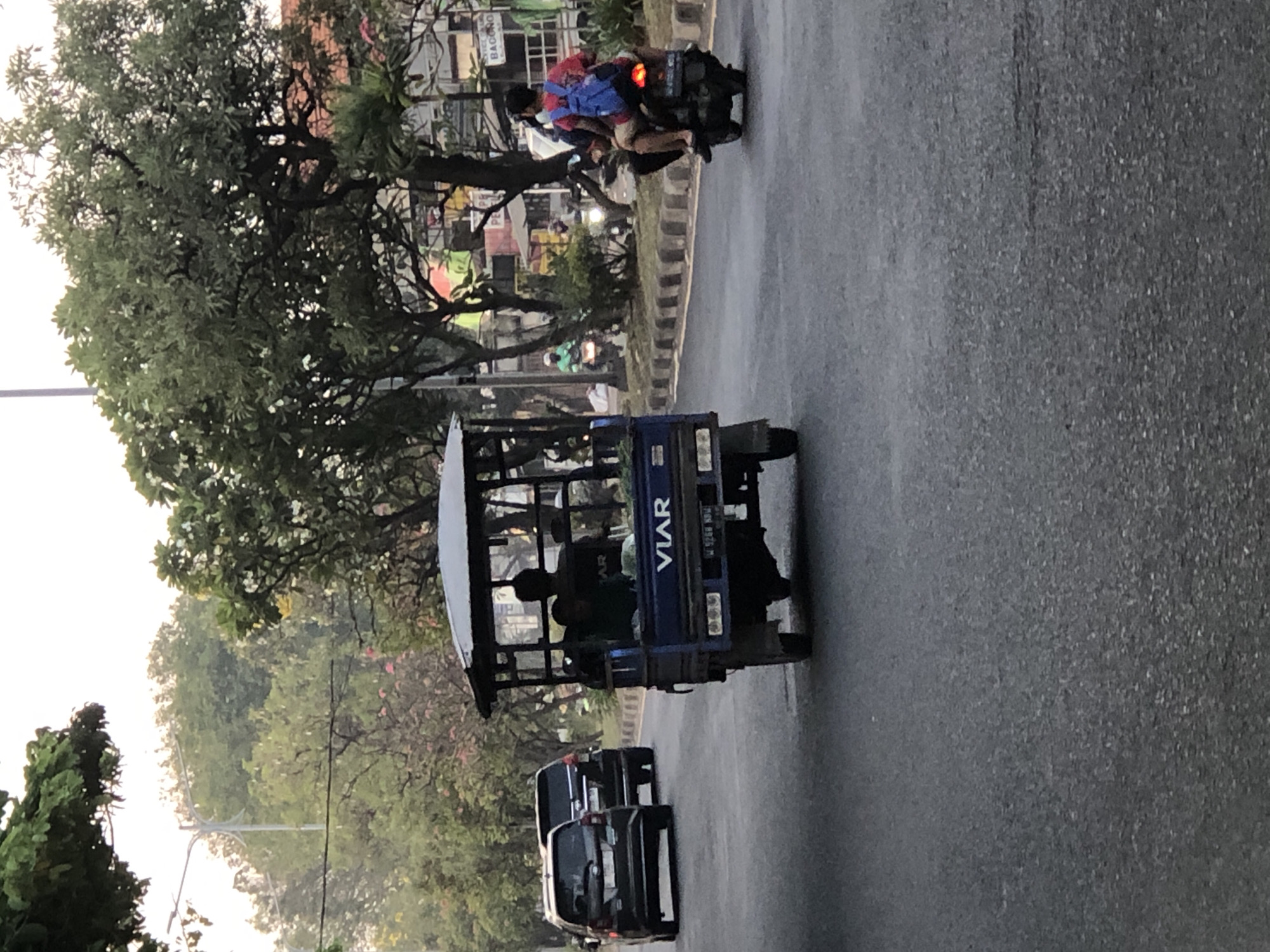 The image depicts a street scene in Sumorame, Indonesia, with several vehicles and people. In the foreground, a blue three-wheeled vehicle, possibly a modified motorcycle or a small truck, is prominent. It has a roof and open sides, with several people visible inside, appearing to be passengers. The vehicle has "VIAR" written on its side. Behind it, two dark-colored cars are driving in the same direction. Further in the background, a motorcycle carrying two people is visible. The rider is wearing a blue backpack, and both individuals appear to be looking forward.
The setting is a paved road with a curb and a grassy median strip. Trees with dense foliage line the sides of the road, some with white flowers. Buildings with signs, though mostly unreadable due to resolution and angle, can be seen in the distance. The lighting suggests it might be late afternoon or early morning, with soft, diffused light. The overall atmosphere is that of a typical street scene in a populated area. There is no clear indication of specific weather conditions, but the road appears slightly wet, suggesting recent rain or humidity. The presence of multiple vehicles and people indicates ongoing daily activity. One visible license plate on the blue vehicle appears to read "W 9266 BDU".