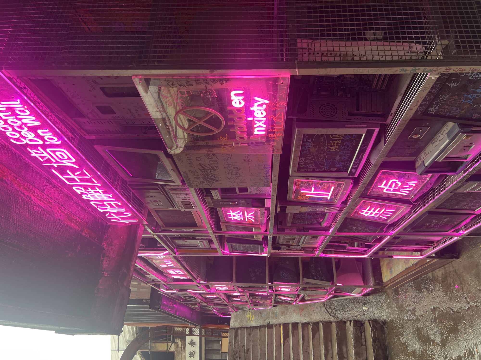 This image captures a section of a street in Yuzhong District, China, illuminated by vibrant pink neon lights. The primary subject is an art installation or storefront display constructed from stacked computer monitors and computer towers, all encased in a metal shelving unit. Pink neon tubing outlines the shelving and highlights several of the monitors, which display various text and symbols.
Prominently featured is a neon sign reading "Forbidden Anxiety" in English, superimposed over a glass panel that has graffiti written on it in Chinese characters. Above this, another neon sign displays Chinese characters that appear to translate to "Code Decomposition Wall." Various Chinese characters are also visible on some of the computer monitors, likely written graffiti or artwork. The overall aesthetic is industrial and edgy, with the old computer equipment juxtaposed against the modern neon signage.
To the left of the display, a set of concrete stairs ascends, with a street sign in the background showing Chinese characters. The ground in front of the installation is wet and reflective, suggesting it may have recently rained or is damp from a nearby water source. The environment appears to be an urban setting, possibly an alleyway or a pedestrian street. No people are visible in the scene.