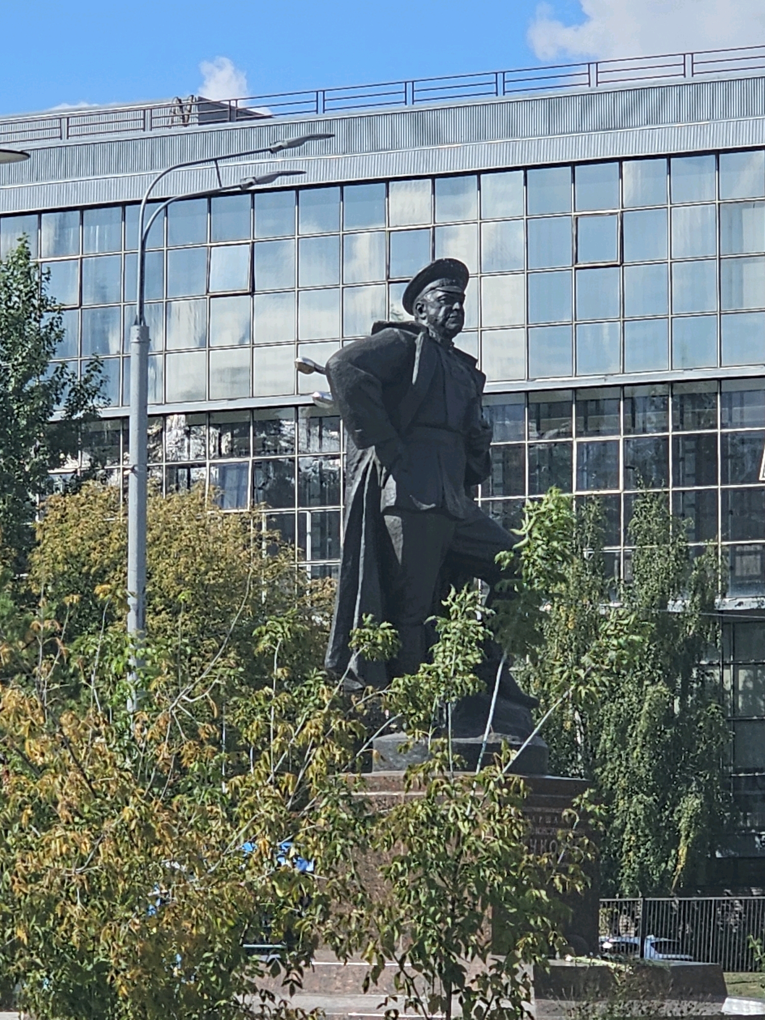 The image shows a statue of a man in a military uniform and hat, standing with his left leg forward. The statue is made of dark material, likely bronze, and is positioned on a large stone pedestal. The pedestal has inscriptions on it, but they are partially obscured by foliage. Behind the statue, there is a modern building with a facade of large glass windows that reflect the sky and surrounding environment. A street lamp with two lights is visible on the left side of the image, in front of the building. Lush green trees and foliage surround the base of the statue and partially obscure the lower part of the building. The sky is blue with some white clouds. The scene appears to be an outdoor public space in a city. This statue is located in Moscow, Russia, and is a monument to Soviet Marshal Grigory Kulik, located on Malaya Gruzinskaya Street.