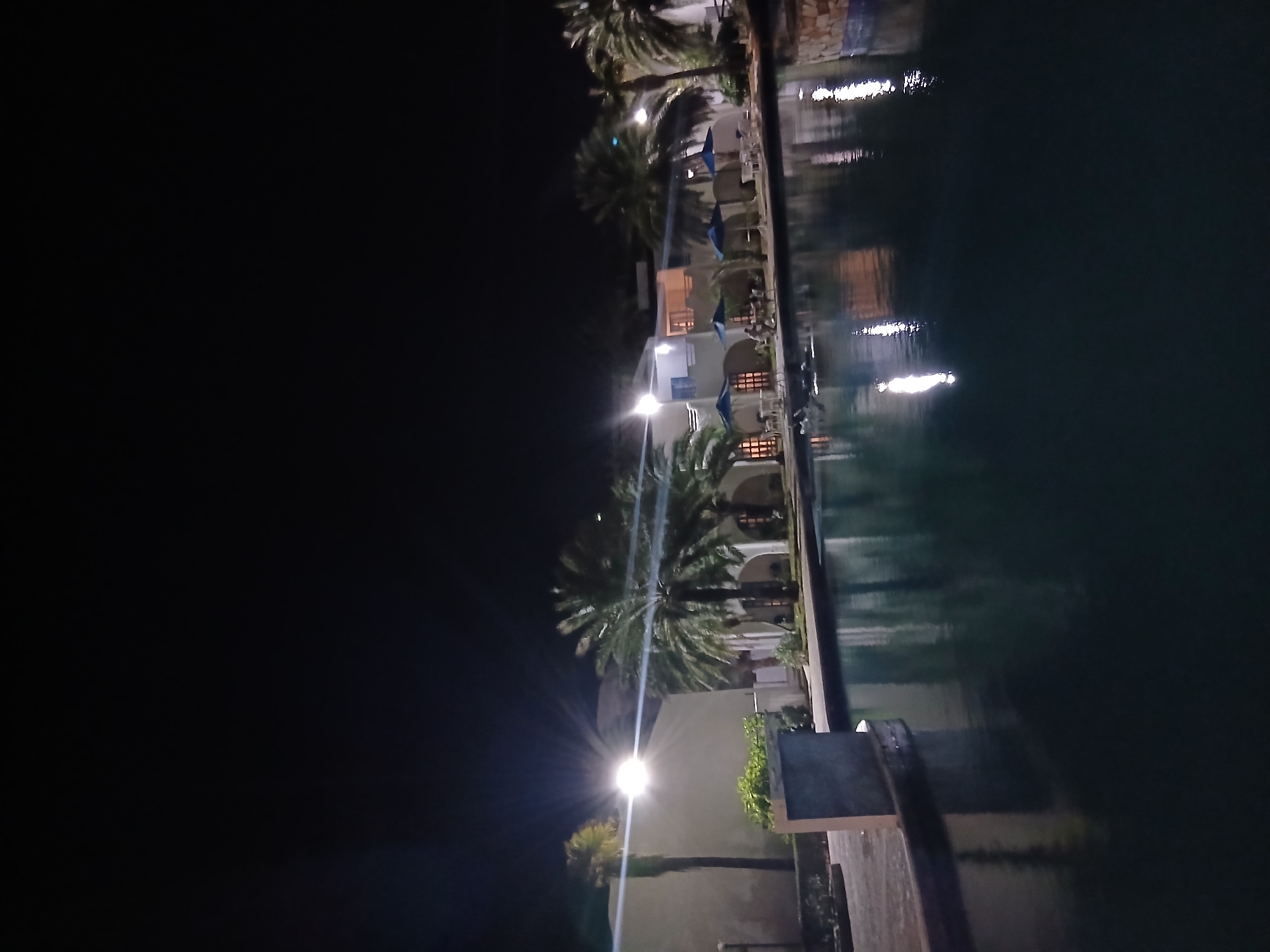 The image depicts an outdoor night scene in Altagracia, Venezuela, likely a resort or hotel complex. A large body of water, possibly a pool or canal, occupies the foreground and midground, reflecting artificial light sources. Along the far bank of the water body, several white or light-colored buildings with arched ground-floor openings are visible. Some building windows are illuminated, casting warm light. Numerous palm trees of varying heights are interspersed among the buildings and along the waterfront.
Multiple bright artificial lights are present, creating starburst lens flares in the upper left and mid-right portions of the image, illuminating parts of the scene. Blue patio umbrellas are set up near the water's edge, accompanied by white tables and chairs. Several indistinct human figures are observed seated at these tables under the umbrellas, engaged in passive activity. The sky is dark, indicating nighttime. The environment is orderly, suggesting a maintained recreational or hospitality area.