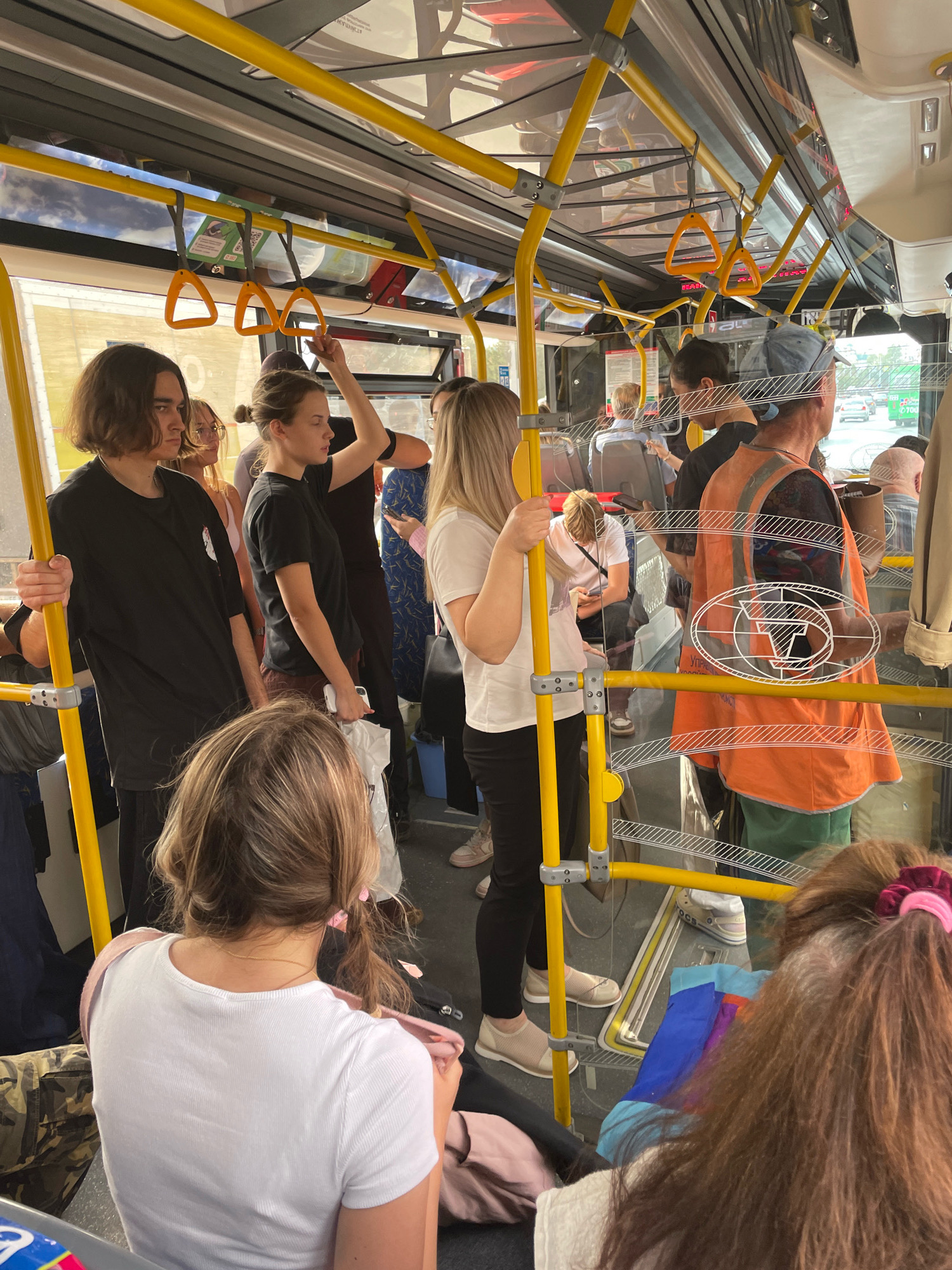 The image shows the interior of a crowded public bus in Omsk, Russia. Several passengers are visible, standing and seated. Yellow handrails and poles are prominent throughout the bus. In the foreground, a person wearing a white t-shirt is seen from the back, and to their right, another person in an orange vest and green pants is also visible. Other passengers, including a man with long hair in a black t-shirt, a woman in a black t-shirt, and others further down the aisle, are present. Outside the windows, a street scene with vehicles and buildings can be observed, indicating the bus is in motion. The overall atmosphere suggests a typical commute on a public transport vehicle.