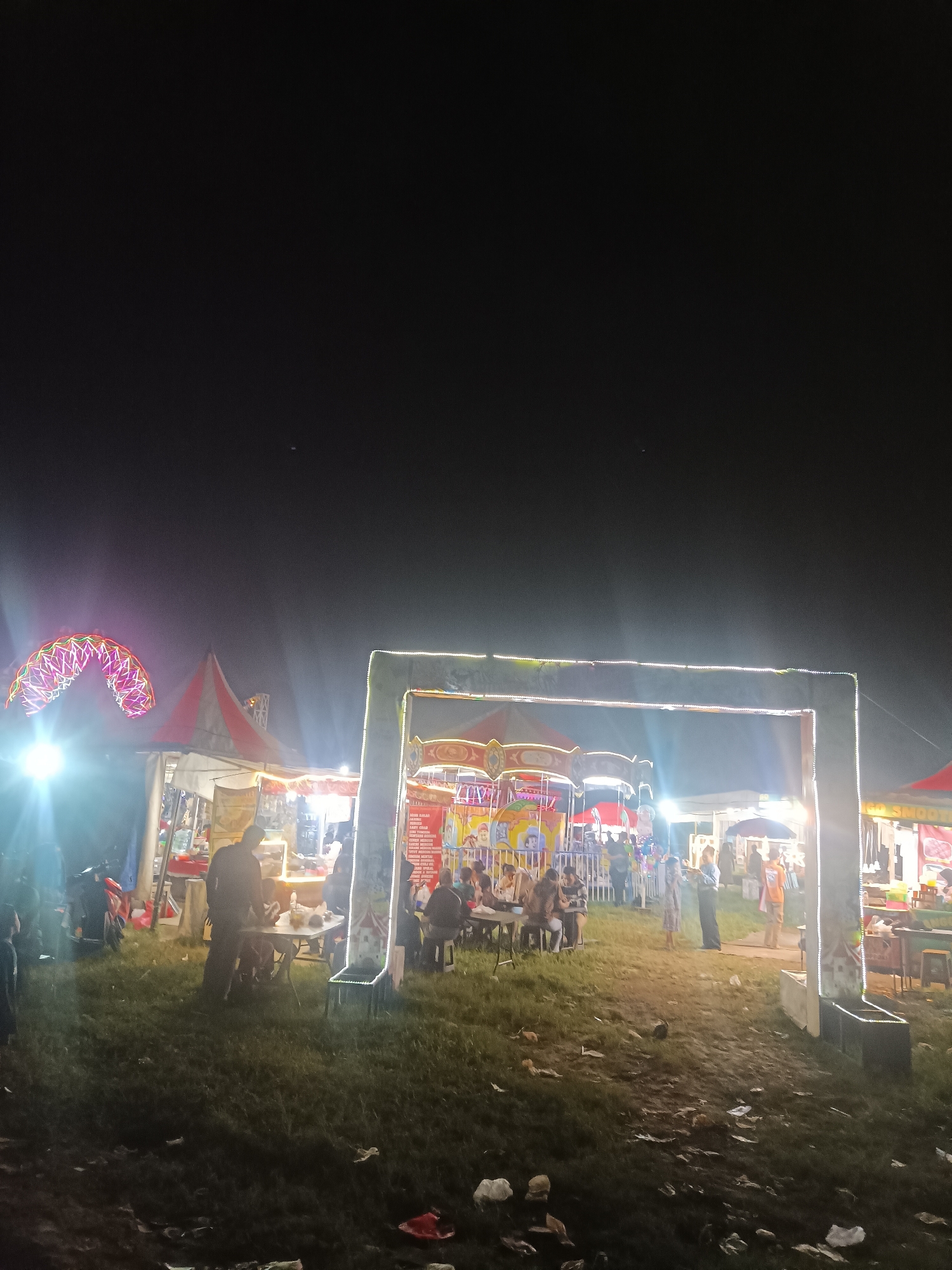 The image depicts a lively night market scene in Depok, Indonesia. A large, illuminated archway serves as an entrance or focal point, adorned with strings of bright lights. Beyond this arch, various stalls and attractions are visible, illuminated by a mix of artificial lighting and the ambient glow of the night sky.
To the left, a Ferris wheel, partially obscured by light flares, is a prominent feature, its colorful lights suggesting a festive atmosphere. Tents with red and white striped roofs house food vendors or game booths, with people gathered around them. In the foreground, a grassy area is scattered with litter, indicating the passage of many visitors.
Several people are visible throughout the scene, either manning the stalls, enjoying the attractions, or socializing. Some are seated at tables, possibly eating or resting. The overall mood appears to be energetic and communal, characteristic of a night market or fair. The time of day is clearly night, with the darkness of the sky contrasting with the vibrant lights of the stalls and attractions.
On a stall to the right, some text is visible, though it is difficult to read clearly. It appears to be a menu or advertisement. The presence of a Ferris wheel, various stalls, and the gathering of people strongly suggest a carnival or night market event. The ground is uneven and covered in grass, with some muddy patches. The weather appears clear, as there are no visible signs of rain or fog.