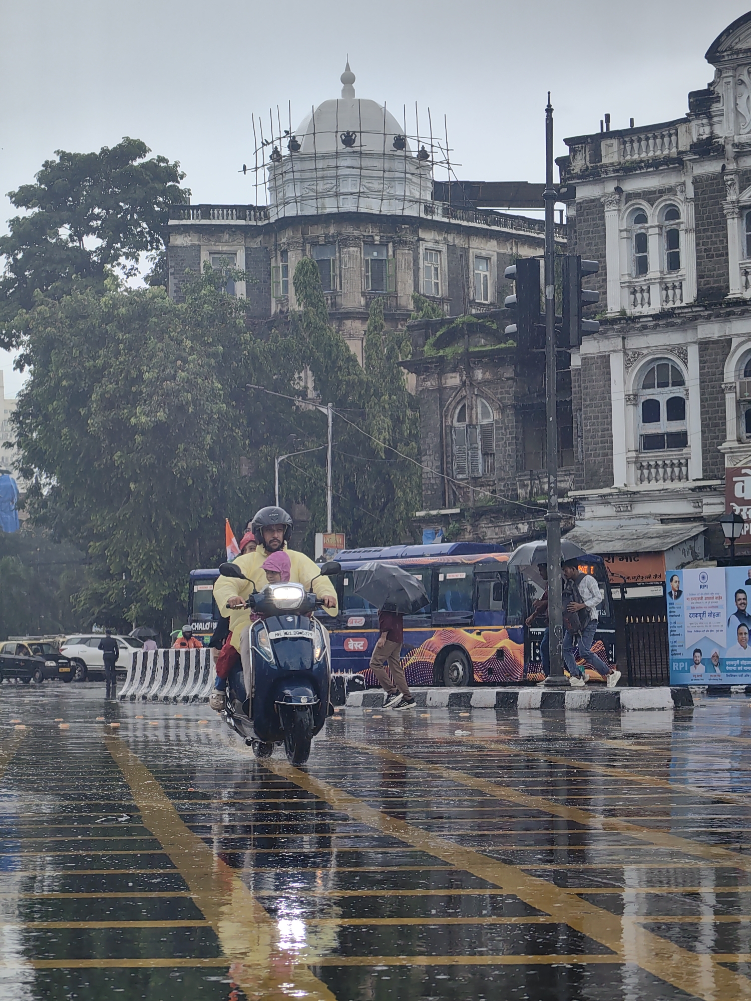 In Mumbai, India, on a rainy day, a man wearing a yellow raincoat and a helmet rides a blue scooter with two passengers. They are traveling on a wet road with yellow and black stripes. In the background, a bus with an orange and yellow design is visible, along with several buildings. One building, appearing to be under construction, is covered in scaffolding. Pedestrians are seen using umbrellas to shield themselves from the rain. The scene depicts a typical street view in Mumbai during monsoon season.