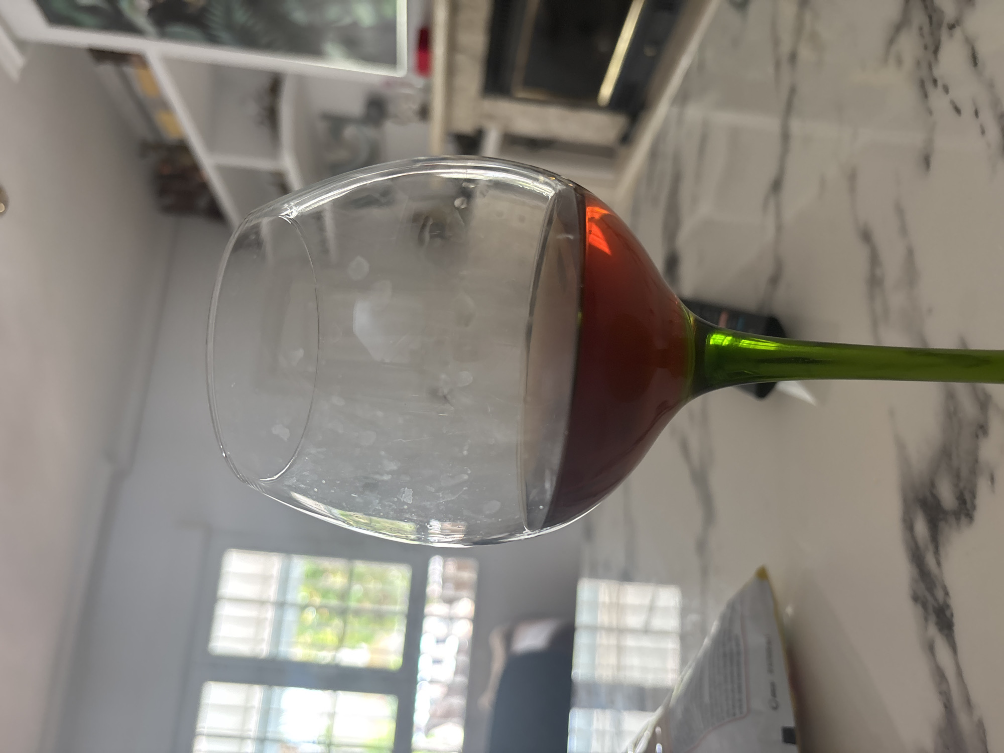 A close-up shot shows a wine glass with a green stem filled with a dark red liquid. The glass has some white residue on its interior surface. The glass sits on a marble-patterned table. In the background, a fireplace with a black insert is visible, flanked by white shelves. Through a window with white blinds, greenery and sunlight can be seen, suggesting an outdoor setting. A white bag with some red and black text is also present on the table to the left of the wine glass. The image appears to be taken indoors, likely in a home, and given the context of the wine glass and the casual setting, it's plausible this is a domestic scene in Orihuela, Spain, during daylight hours.