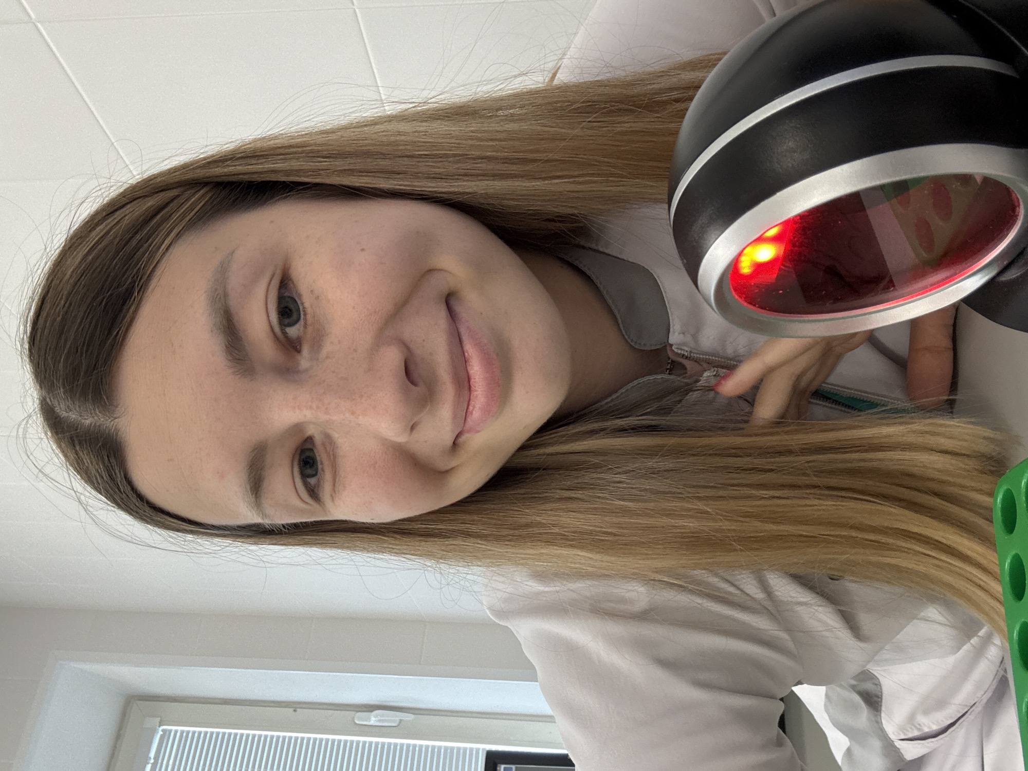 A young Caucasian woman with blonde hair and fair skin is pictured in a close-up selfie. She is wearing a white lab coat with grey trim. Her left hand is resting on her chest. In the foreground, to her right, is a black device with a red light emanating from its lens. The background consists of a white tiled wall and a window with horizontal blinds. The scene appears to be indoors, possibly in a medical or laboratory setting. There are no discernible landmarks to suggest a specific location in Moscow, Russia.
