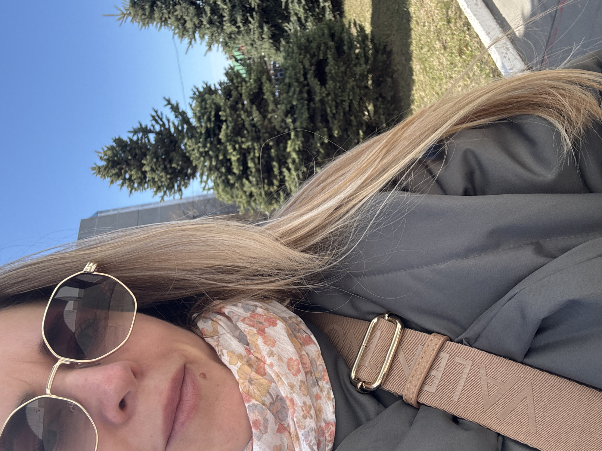 The image presents a close-up view of a person, likely female, positioned outdoors on what appears to be a sunny day. The individual is wearing sunglasses with gold frames and a floral-patterned scarf. A gray-green jacket is also visible, with a tan-colored strap bearing the text "VALENT" crossing the body. In the background, there are coniferous trees and a clear blue sky. A segment of a building and some grass are also seen. The setting appears to be an urban environment, possibly within the city of Chaikovsky, Russia, indicated by the user's request.