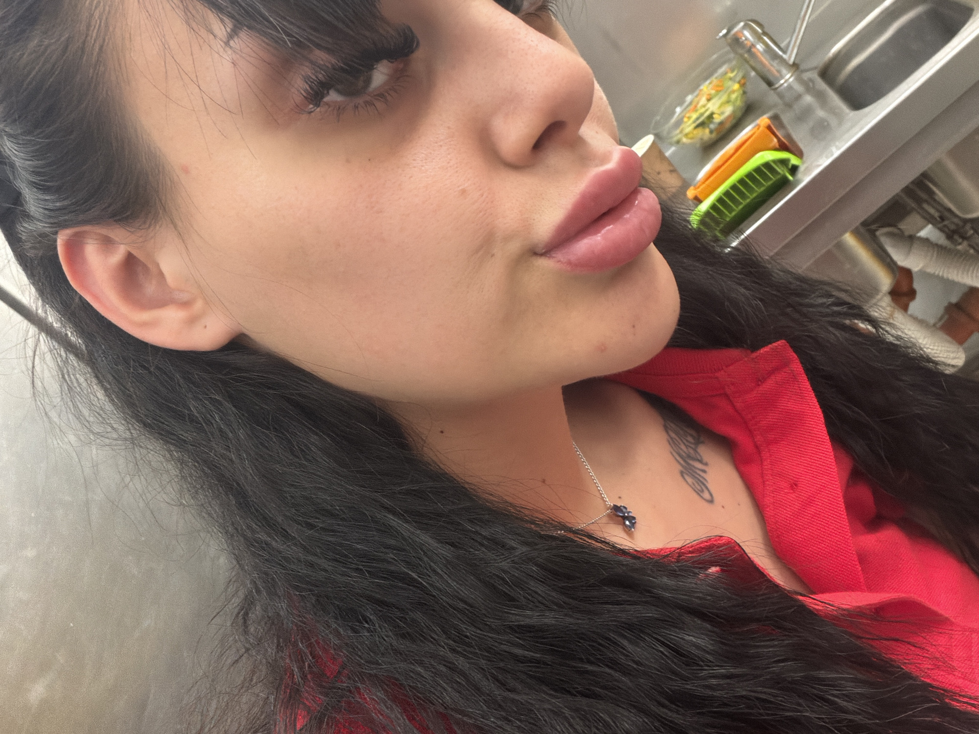 The image is a close-up, angled selfie of a young woman with long, dark hair and bangs. She is wearing a red top and a silver necklace with a dark blue pendant. Her lips are full and painted with a pinkish-red lipstick. A tattoo is visible on her collarbone area. In the background, a stainless steel sink is visible with a faucet, a small green and orange food divider, and a clear container with a salad. The scene appears to be in a kitchen or food preparation area. There is no indication of the city of Plovdiv, Bulgaria.