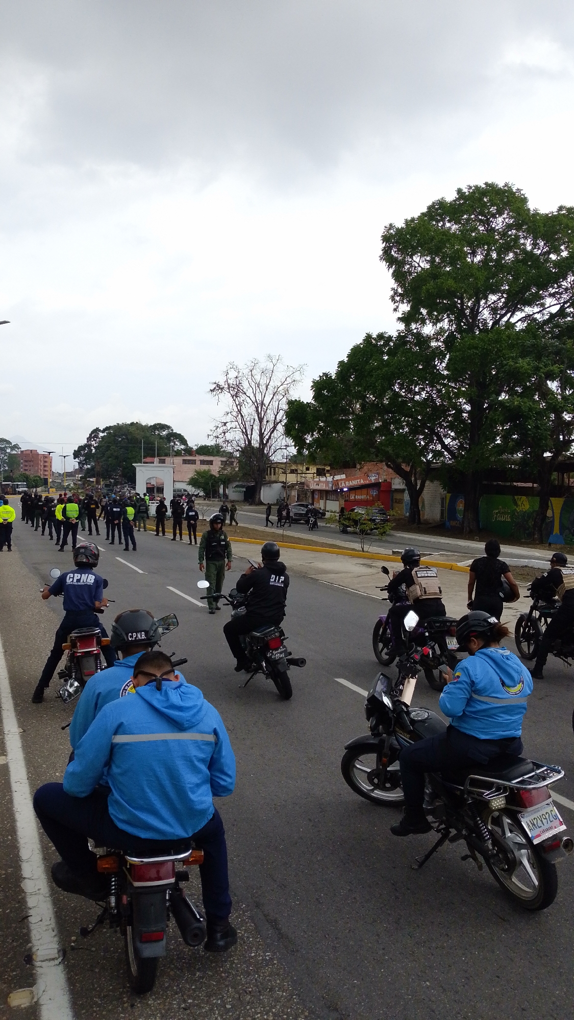 The image depicts a street scene in Tinaquillo, Venezuela, with multiple individuals, primarily law enforcement officers. A group of police officers are standing in a line, seemingly monitoring the area. Several police officers are also on motorcycles, some displaying license plates. The officers on motorcycles are wearing various colored uniforms including dark blue, light blue, and black. The road appears to be paved and is divided by lane markings. The sky is overcast. There are buildings and trees visible in the background.