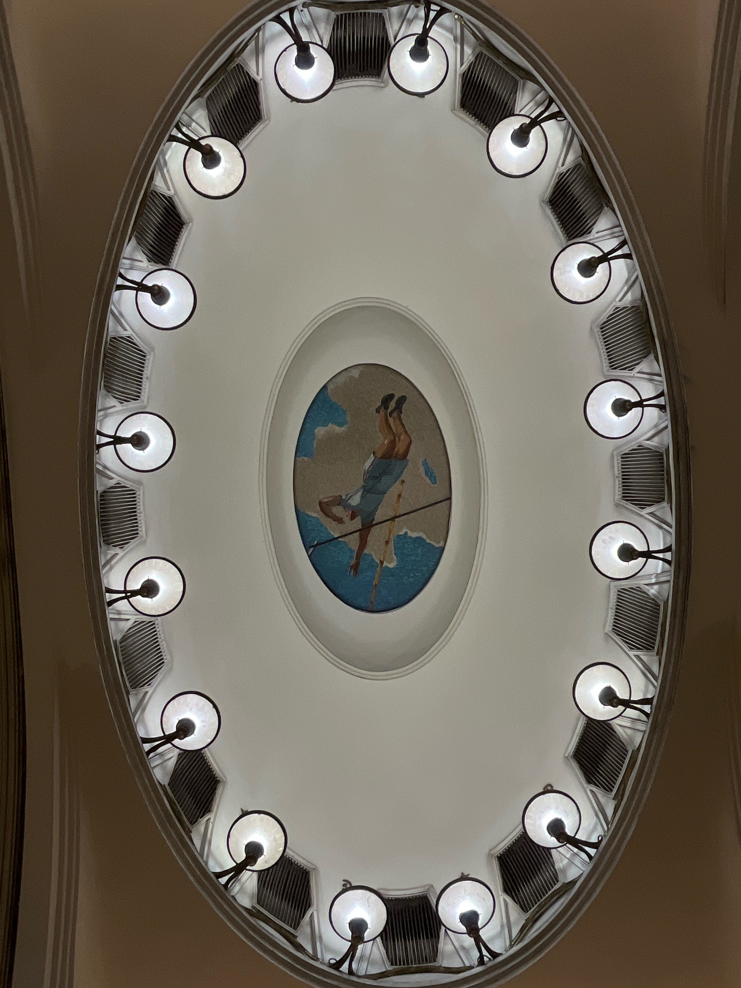 The image shows an oval ceiling with a mosaic in the center depicting a person in mid-air. The mosaic is surrounded by a series of lights and decorative grilles. The lighting is dim, casting a soft glow on the ceiling. The overall impression is one of grandeur and artistic expression. This image was taken in the Novokuznetskaya metro station in Moscow, Russia.