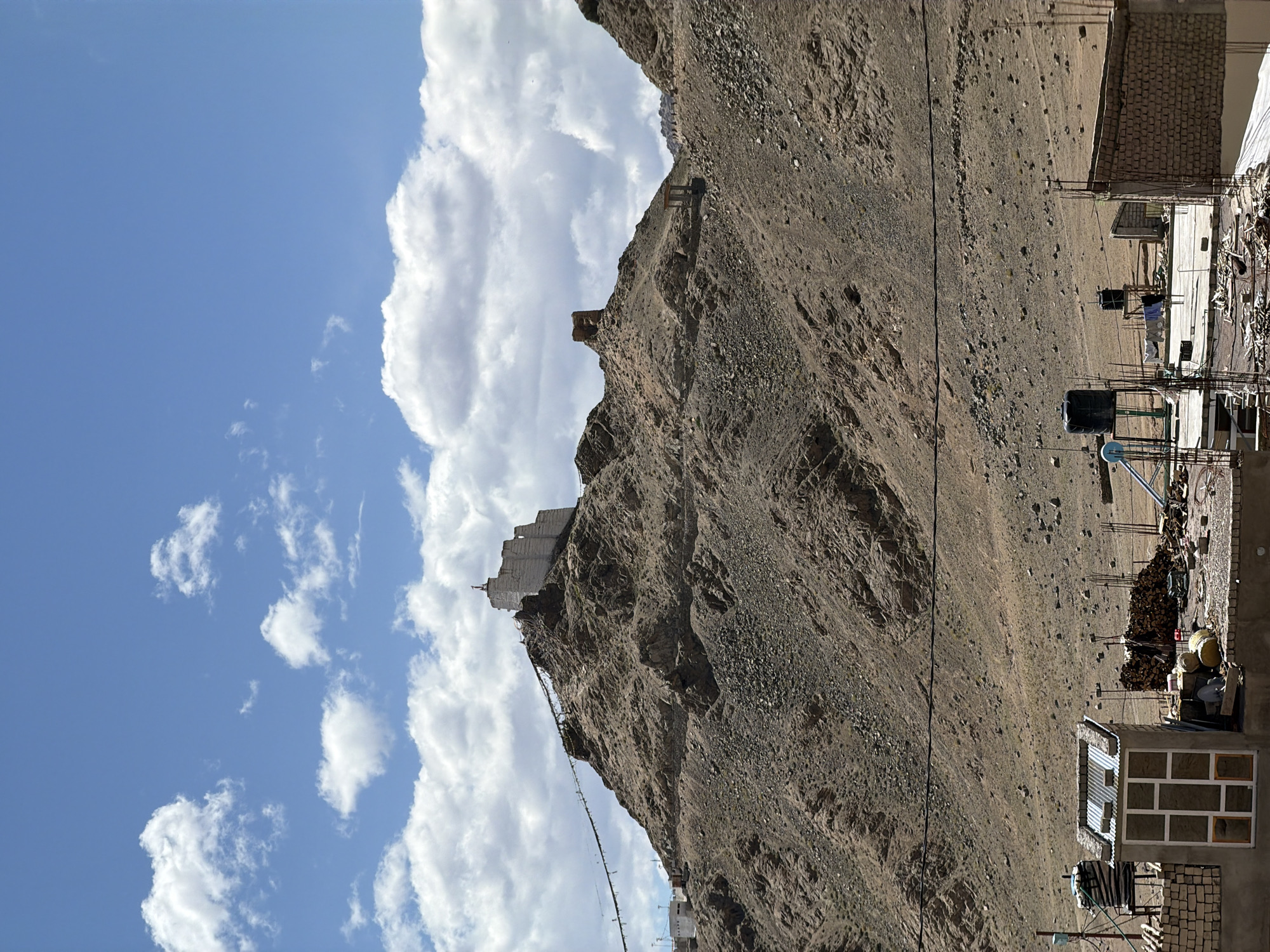 The image shows a view of Leh, India, with the prominent feature being a large white building or monastery perched atop a rocky, arid mountain. The building, likely Leh Palace or a similar historical structure, is white and has a distinctive tiered architecture. In front of it, a string of prayer flags stretches across a ridge leading up to the structure. The mountain itself is covered in loose rocks and sparse vegetation, typical of the Himalayan landscape. The sky above is a bright blue, dotted with scattered white cumulus clouds.
In the foreground, the scene shifts to a more developed area with several buildings and structures. A small, multi-paned windowed structure with a corrugated metal roof is visible on the left. Further back, various construction materials like piles of wood and rebar suggest ongoing building or repair work. Water tanks, one large black one and a smaller one, are also present. The overall impression is a contrast between the ancient, majestic structures on the mountains and the more utilitarian, developing urban landscape at the base. The lighting suggests it is daytime, likely late morning or early afternoon, with the sun casting distinct shadows.