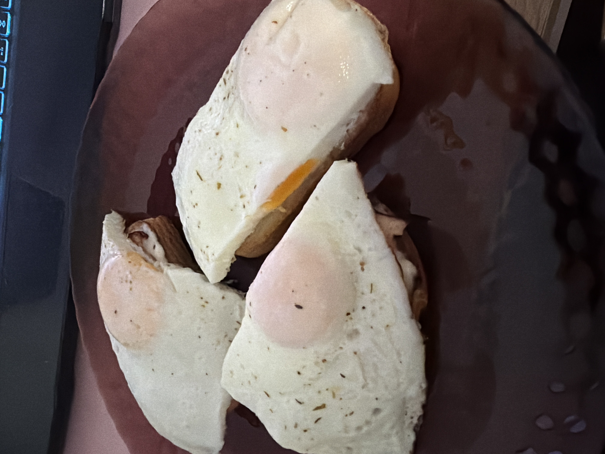 Three fried eggs are placed on top of toasted bread slices, which are arranged on a dark red plate. The eggs appear to be sunny-side up, with one egg's yolk partially visible and runny. The toast slices are slightly browned, and there are herbs sprinkled on top of the eggs. The plate is positioned on a pink surface, with a dark object, possibly a laptop keyboard, visible in the background. The scene appears to be indoors, with no specific details indicating a location in Chaikovsky, Russia.