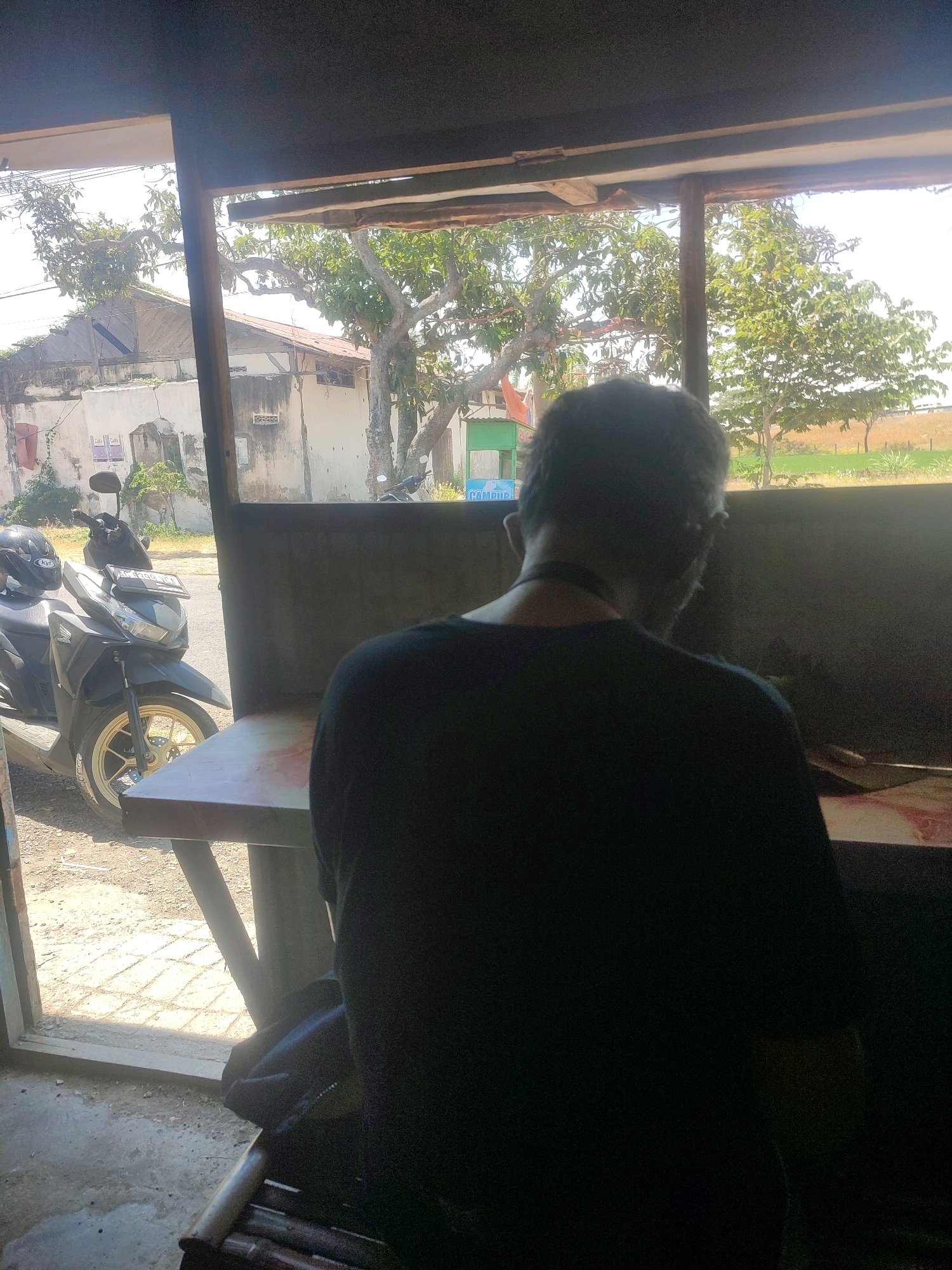 An individual with dark hair and a dark shirt is seated indoors, facing away from the camera, looking out a window or open doorway. The person is positioned in front of a table and a dark wall. Outside, a scooter is parked on a dirt road. Further in the background, there are trees, buildings, and a field under a bright sky. The scene appears to be a roadside stall or eatery. The location is Putren, Indonesia.