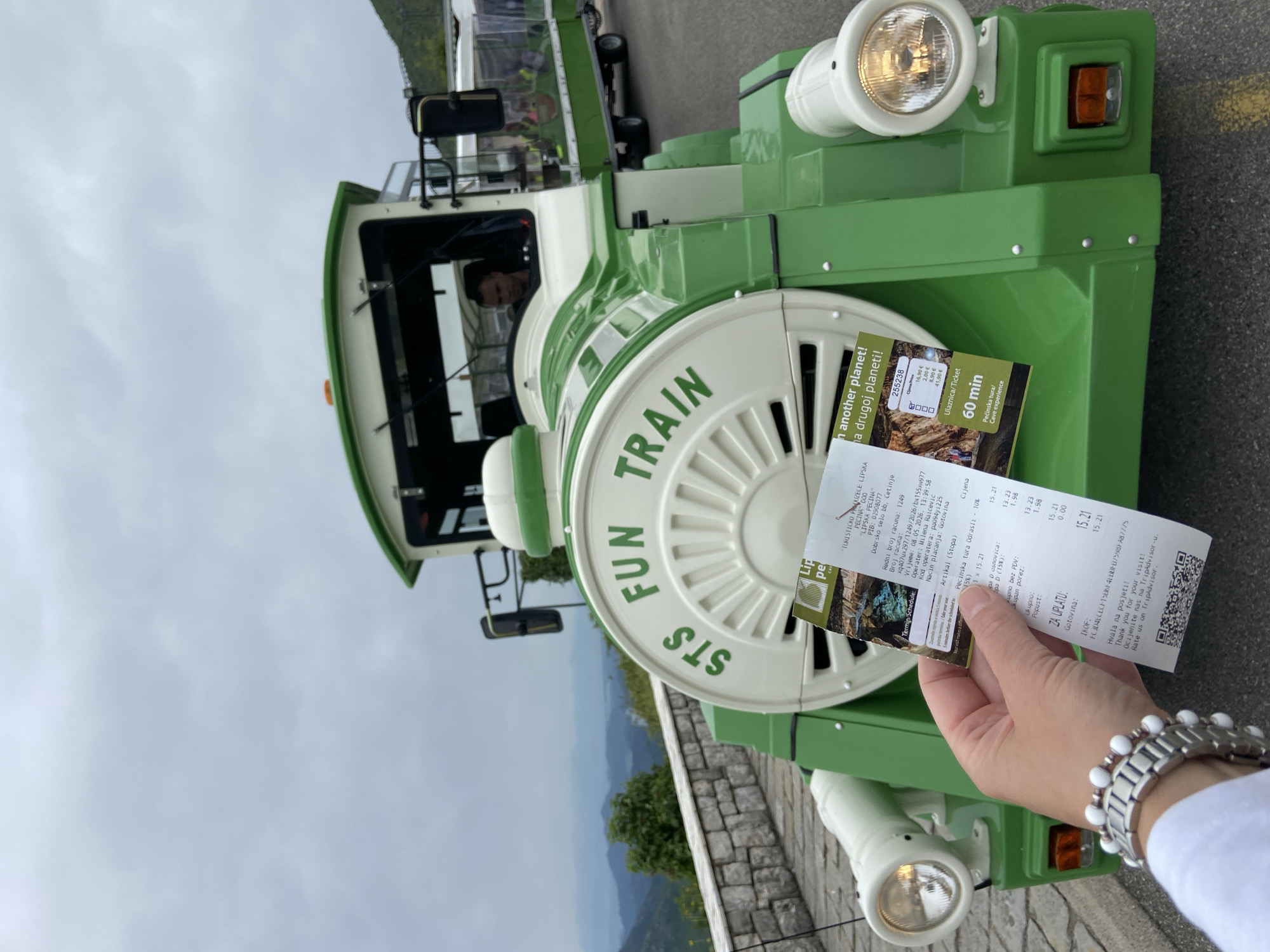 The image shows a green and white "Fun Train STS" tourist train, likely in Lipa Dobrska, Montenegro, indicated by the ticket. A hand wearing a silver and white beaded watch holds a ticket and brochure in front of the train's large circular front. The ticket is from "Lip pe" for "Pecinska tura Odraslı 10% Pecinska tura/Cave experience 60 min." The terrain behind includes a grey cloudy sky and trees visible in the distance.