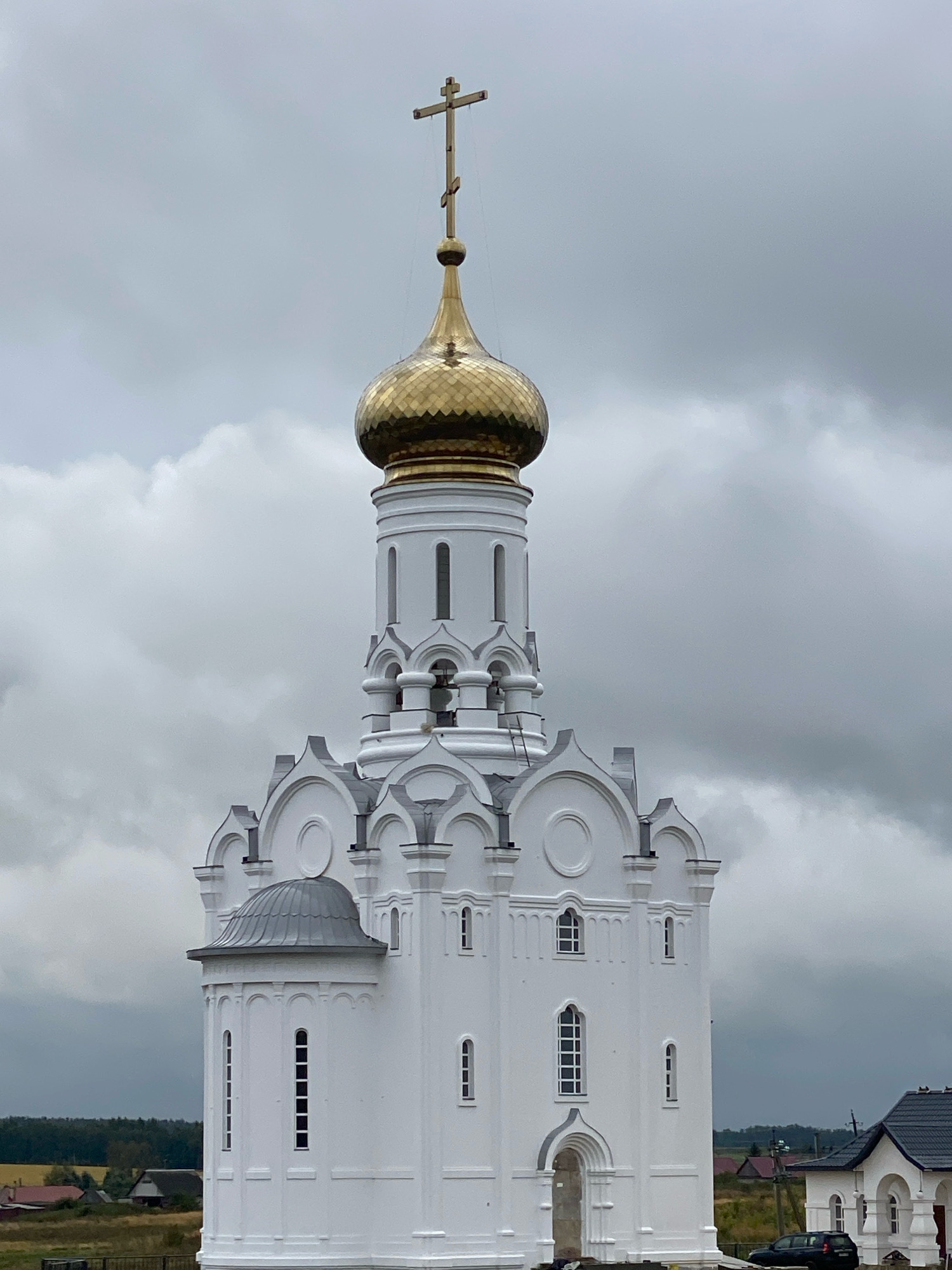 The image shows a white Orthodox church with a golden dome and a cross on top, set against a cloudy sky. The church has several windows of various shapes and sizes, and intricate architectural details. To the right of the church, a smaller white building with a dark roof is visible, and a black car is parked in front of it. The background shows a rural landscape with fields and trees, and some houses. The location is identified as Беломестная Криуша, Russia. The scene appears to be peaceful and serene, with no people or animals present. The weather is overcast, contributing to a somber mood.