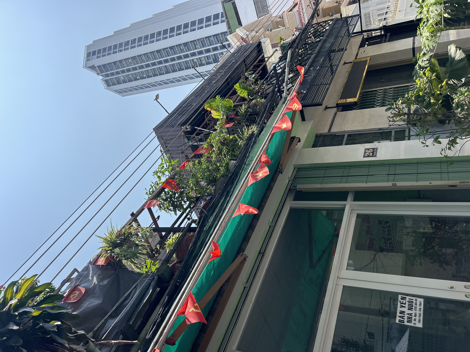 Captured from a low angle, the image showcases a narrow building facade in what appears to be a densely populated urban area. The building is adorned with small Vietnamese flags, adding a vibrant touch to the scene. A makeshift scaffolding or balcony structure is visible, embellished with a variety of potted plants. Overhead, a modern high-rise building looms in the background, contrasting with the older architecture of the foreground. The environment appears sunny, with clear blue skies visible above the structures. The scene is cluttered with wires, indicative of a densely populated urban setting. The building address "26" is visible near the front entrance, and a sign for "BÁN YẾN NHÀ NUÔI" (Bird's Nest House for Sale) is pasted to the building. Based on architectural style and signs, the location is likely in Bắc Nha Trang, Vietnam.