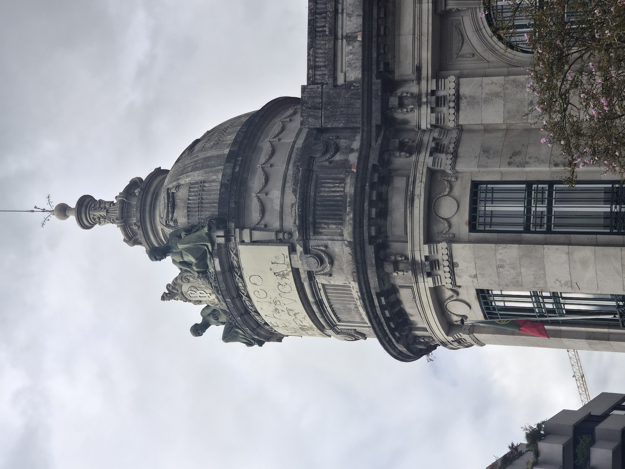 This is a low-angle, full shot of the Banco de Portugal building in Braga, Portugal, under a cloudy sky. The image focuses on the ornate dome and upper facade of the building, which is constructed of grey stone with intricate architectural details. Two seated statues, weathered and dark, adorn the top of the dome, flanking a shield with the letters "BP" inscribed. Below them, a rectangular plaque is visible with raised lettering that reads "BANCO DE PORTUGAL". The building features large, segmented windows with dark green bars, and a Portuguese flag hangs from a flagpole to the left of a prominent window. To the left of the Banco de Portugal building, a modern grey building with potted plants is partially visible. A construction crane is visible in the background between the two buildings. Pink flowering branches are in the foreground on the right side of the image.