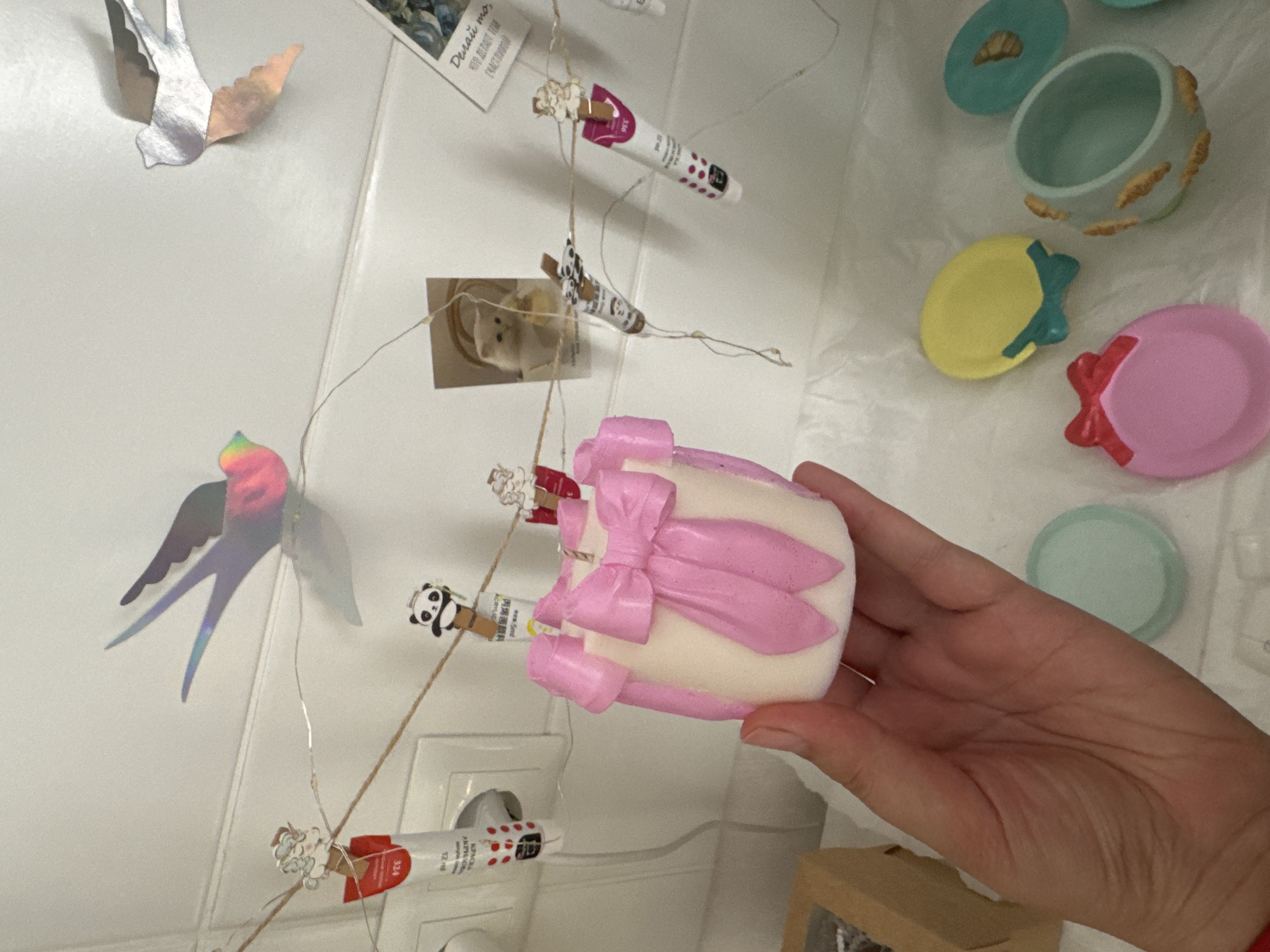 A hand holds a pink and white candle shaped like a gift with a bow. The candle is on a white surface next to several small, colorful dishes and a light string with various small objects clipped to it, including tiny tubes and paper cutouts of birds, a panda, and a cat. The background is a white, tiled wall.