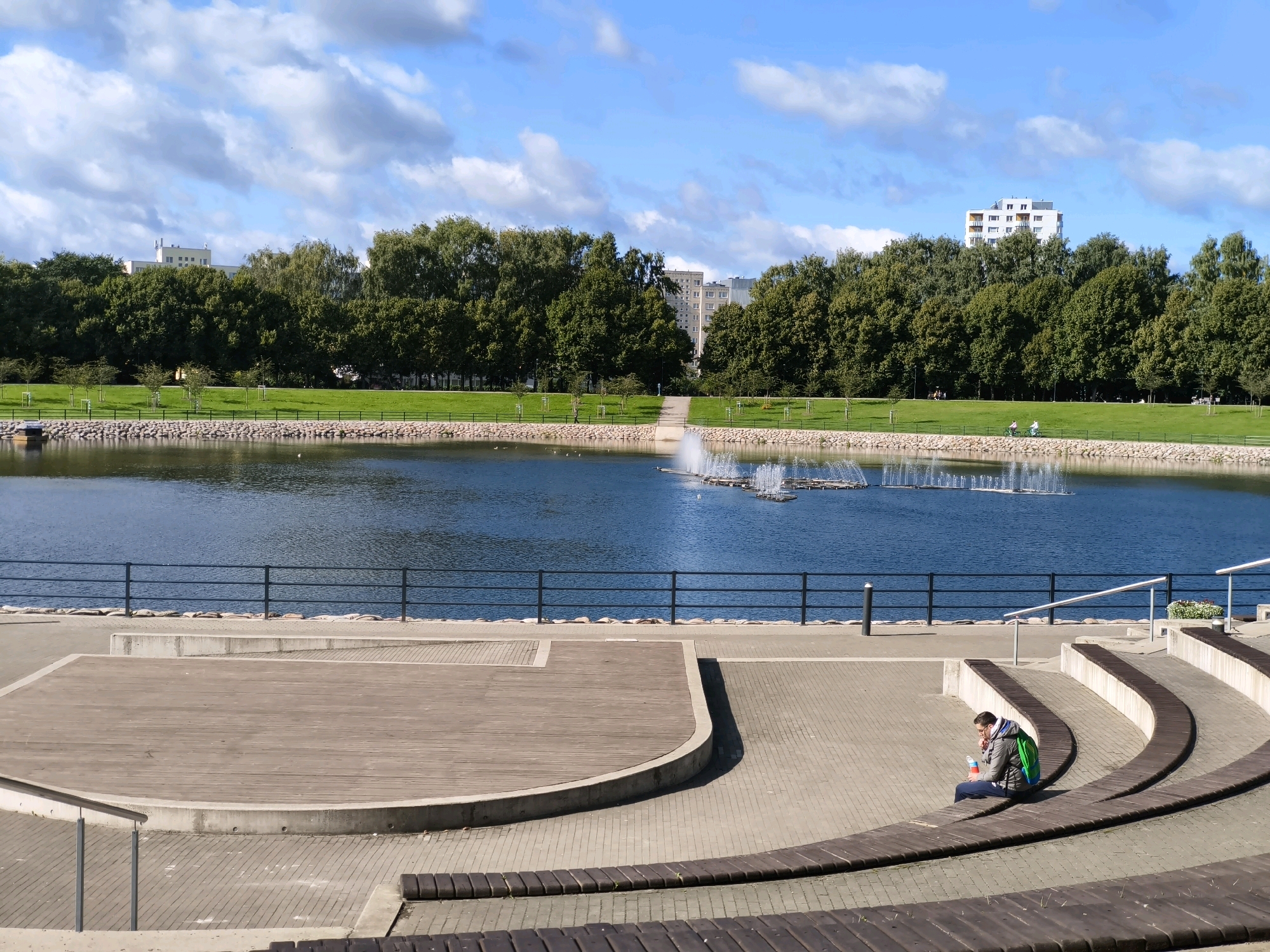 This is a daytime, eye-level, medium shot photograph of a park area in Tallinn, Estonia. In the foreground, an amphitheater-like seating area made of concrete and wood curves inward. A person wearing a green backpack and a grey jacket is seated on the wooden steps, looking down at a white container and a red object in their hands. To the left, a circular wooden platform is visible. Beyond the seating area, a black metal fence separates the paved area from a large blue lake. In the lake, a series of fountains are in operation, creating white spray. The far bank of the lake is lined with a neatly mown green lawn, with a line of trees behind it. In the distance, several modern, multi-story buildings are visible peeking out from behind the trees. The sky is blue with scattered white clouds.