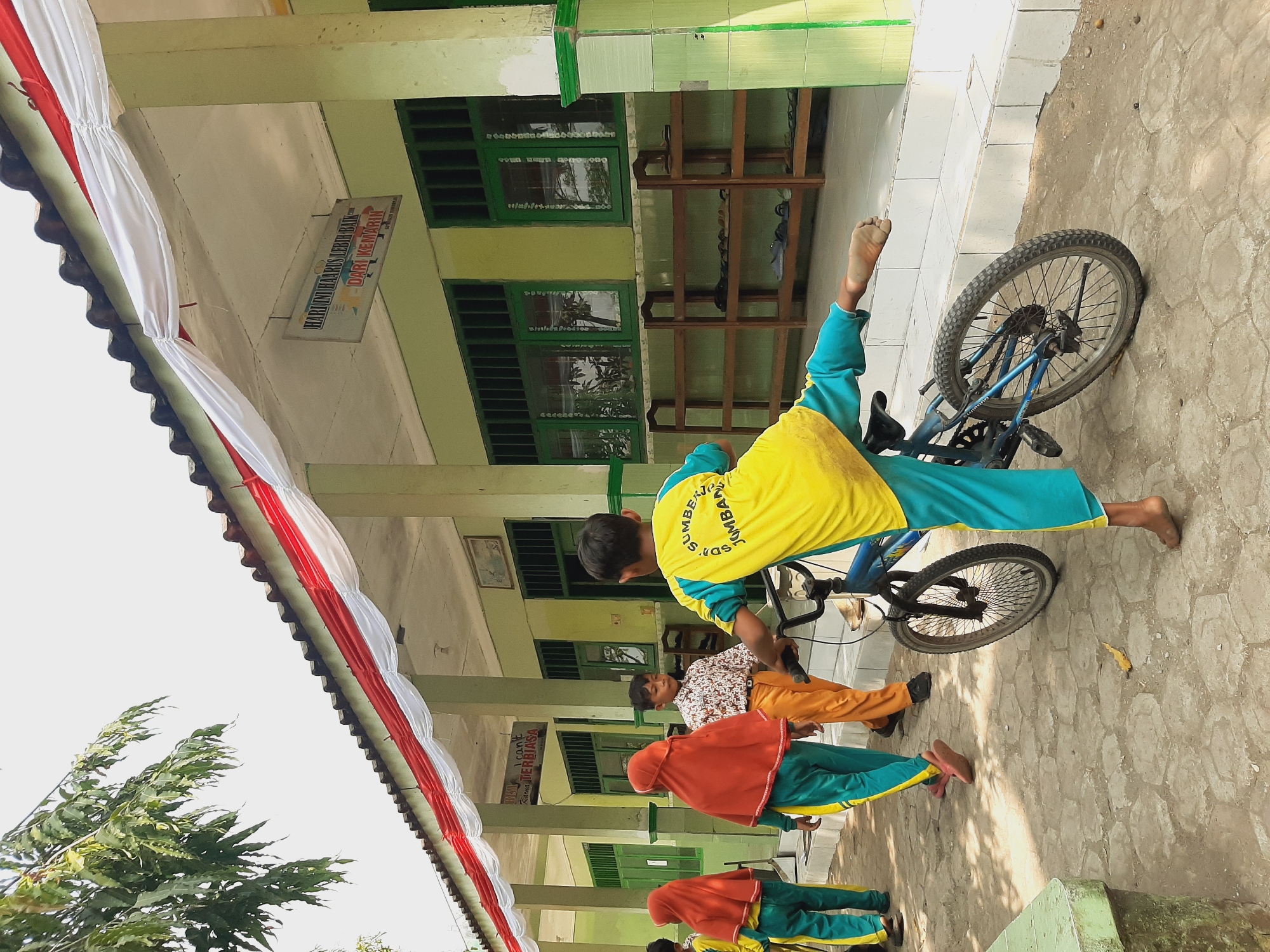 In front of a school building in Sumberjo, Indonesia, a group of children are gathered. A boy in a yellow and blue uniform is standing next to a blue bicycle, with one leg extended behind him as if he is about to kick off or balance. Other children, also in uniform, are walking past. One girl wears an orange headscarf and a matching jumpsuit, while another wears a yellow and green uniform. A boy in a patterned shirt and orange pants walks in the opposite direction. The school building is painted green with white and red banners draped along the roofline, and it has green-barred windows. A shoe rack is visible along the exterior wall. The ground is paved with uneven stone tiles.