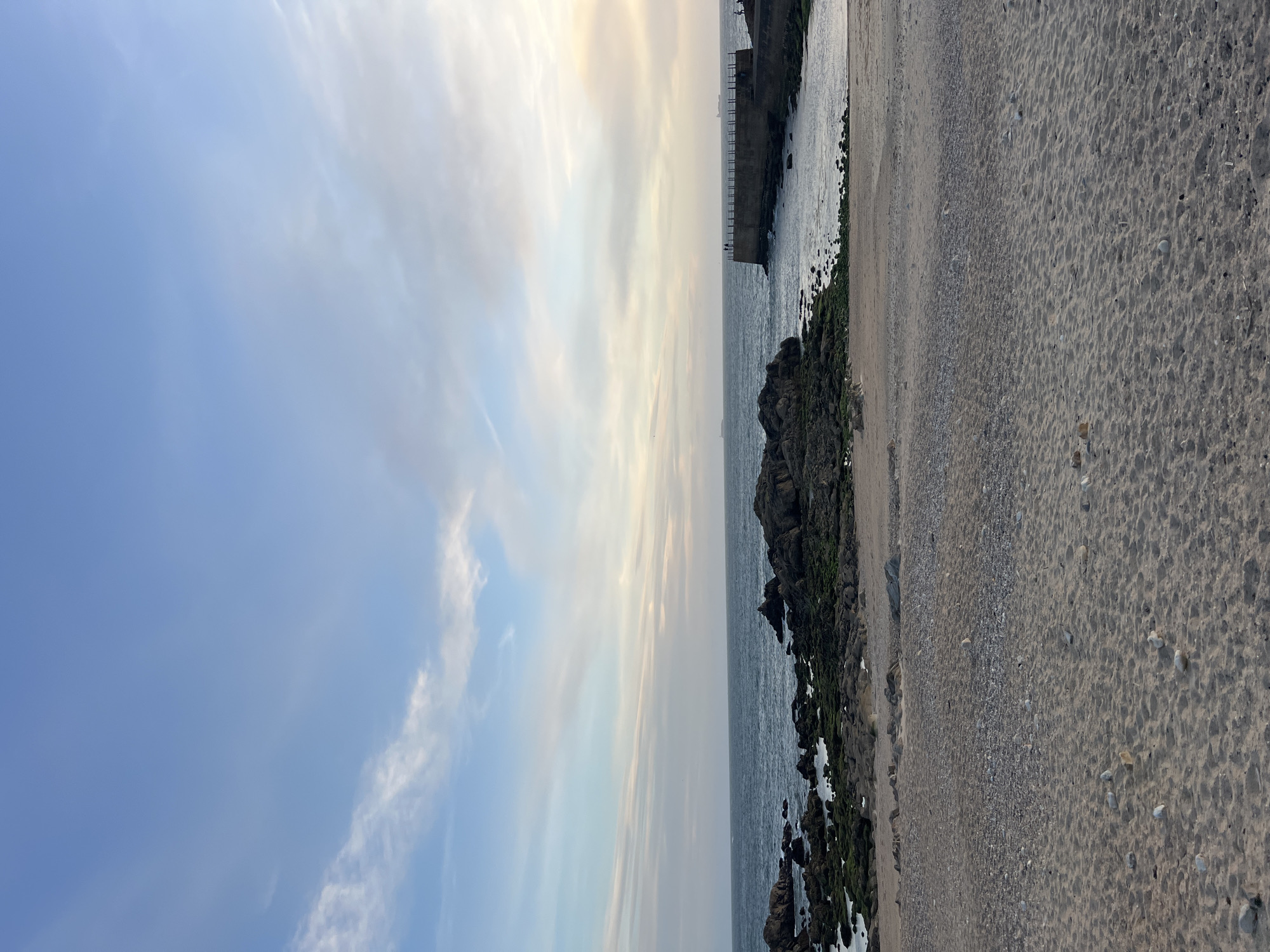 The scene depicts a beach in Porto, Portugal. The view is oriented vertically, with the sky on the left and the sandy beach on the right. The sky is a mix of light blue and white clouds, suggesting either early morning or late afternoon. To the right of the sky is the sea, with rocky outcrops covered in greenish algae along the shore. Further into the sea, a concrete structure, possibly a pier or breakwater, extends into the water. The beach is composed of sand and small pebbles, with visible patterns of footprints or waves. The scene is quiet, with no people immediately visible.