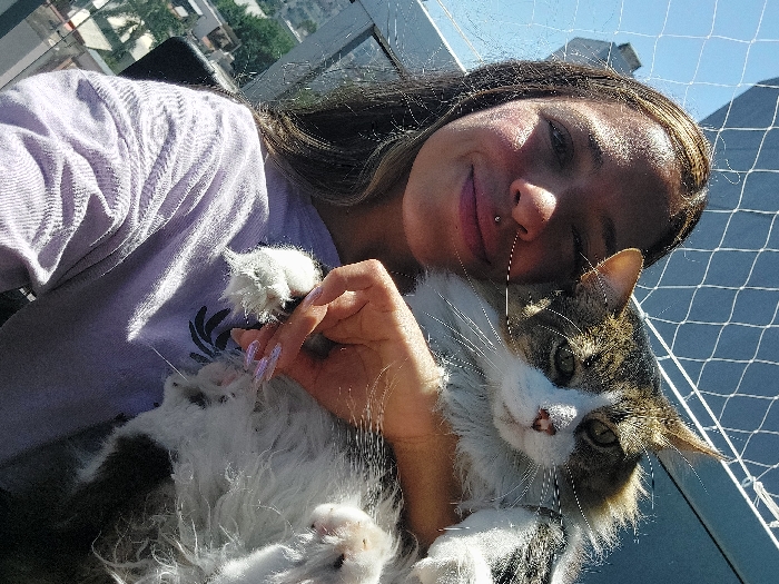 A young woman is depicted in a close-up, slightly tilted image, affectionately holding a fluffy cat outdoors, likely on a balcony in Buenos Aires, Argentina. The woman, with long dark hair, sports a gentle smile and a lip piercing. She wears a light purple long-sleeved shirt, and her hand, adorned with long, iridescent acrylic nails, is visible near the cat's paw.
The cat, a medium-sized, long-haired feline, features distinctive tabby markings in brown, black, and white, with a prominent white chest and muzzle. It appears calm and content, comfortably nestled in the woman's arms. Both subjects are looking towards the camera, conveying a sense of companionship.
The background shows an urban landscape under a bright, clear sky, indicative of daytime. Distant buildings and some greenery are visible. A white safety net, common on balconies, extends across the upper right portion of the frame, providing a safety barrier for the outdoor setting. Strong sunlight casts shadows on the woman's face, emphasizing the bright conditions. The overall mood of the image is one of affection and relaxed companionship. No text is visible in the image.
