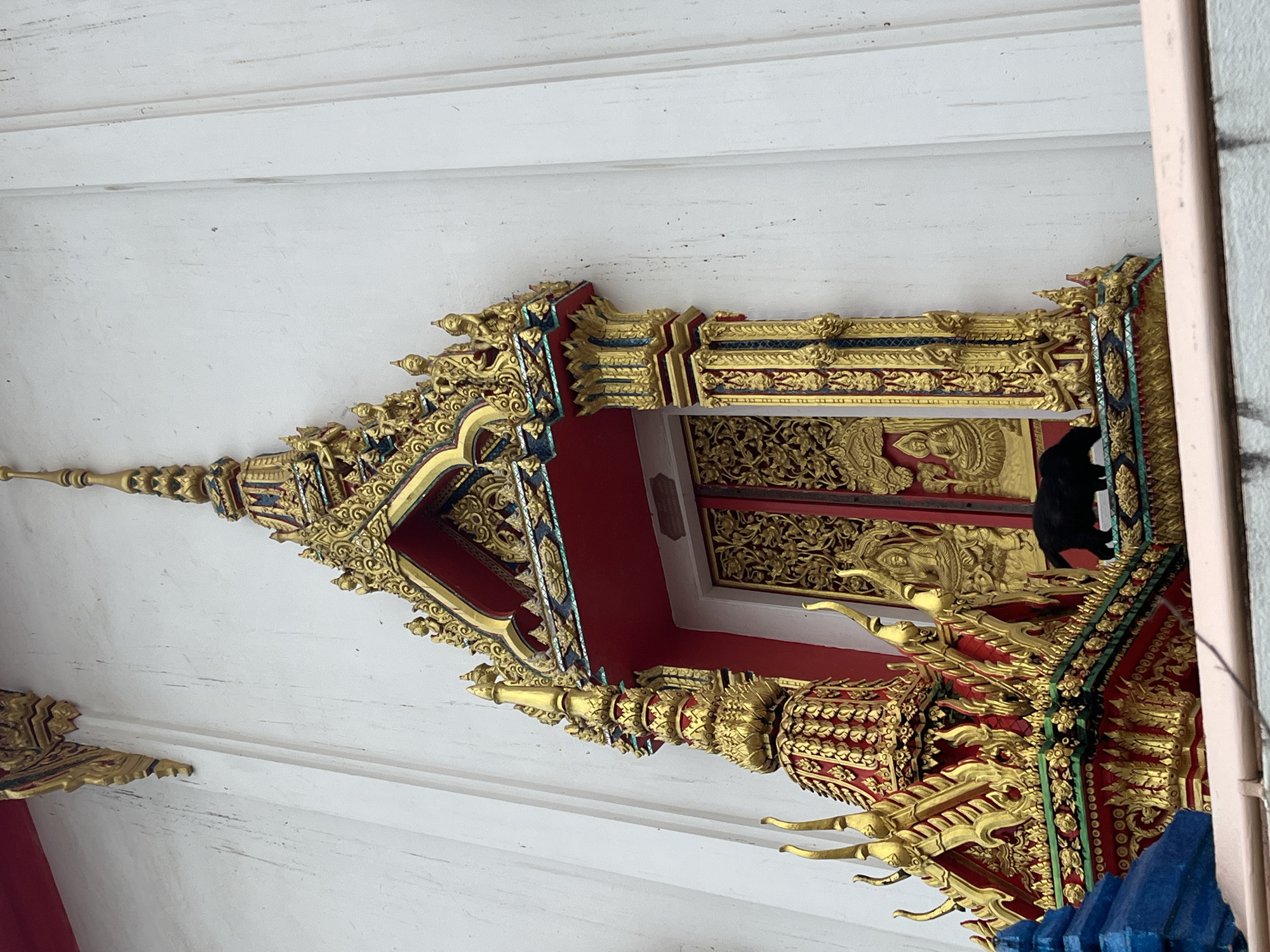 A black cat walks on a decorative, ornate gold and red ledge below a closed, gilded double door set into a white wall. The door is intricately carved and features depictions of figures. The ledge is part of a larger, elaborately decorated architectural element with multiple tiers and spires, typical of Thai temple architecture. The wall appears to be made of stucco and shows some signs of weathering or dirt. The overall scene suggests a temple or religious building in Si Sunthon, Thailand.