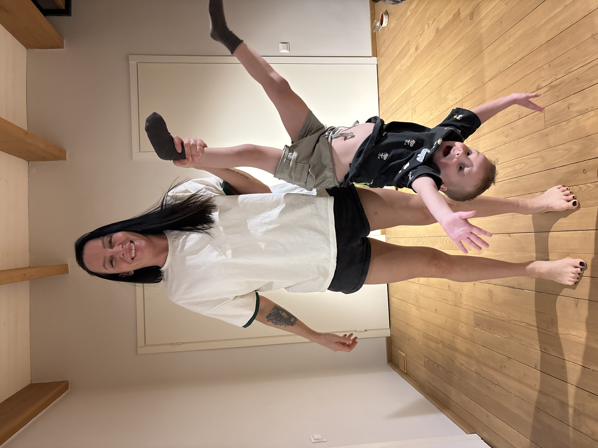 A woman is lying on her back on a wooden floor, holding a boy in the air with her feet. The boy is lying horizontally across her legs, with his arms outstretched. The woman is smiling and looking at the camera. The boy is also smiling with his mouth open, looking at the camera with his legs apart. The woman is wearing a white t-shirt, and black shorts. The boy is wearing a black t-shirt and tan shorts. The background includes a white door and tan walls. The location of the photo is a room with wooden flooring and it's unlikely to be in Chaikovsky, Russia, as it is a private moment.