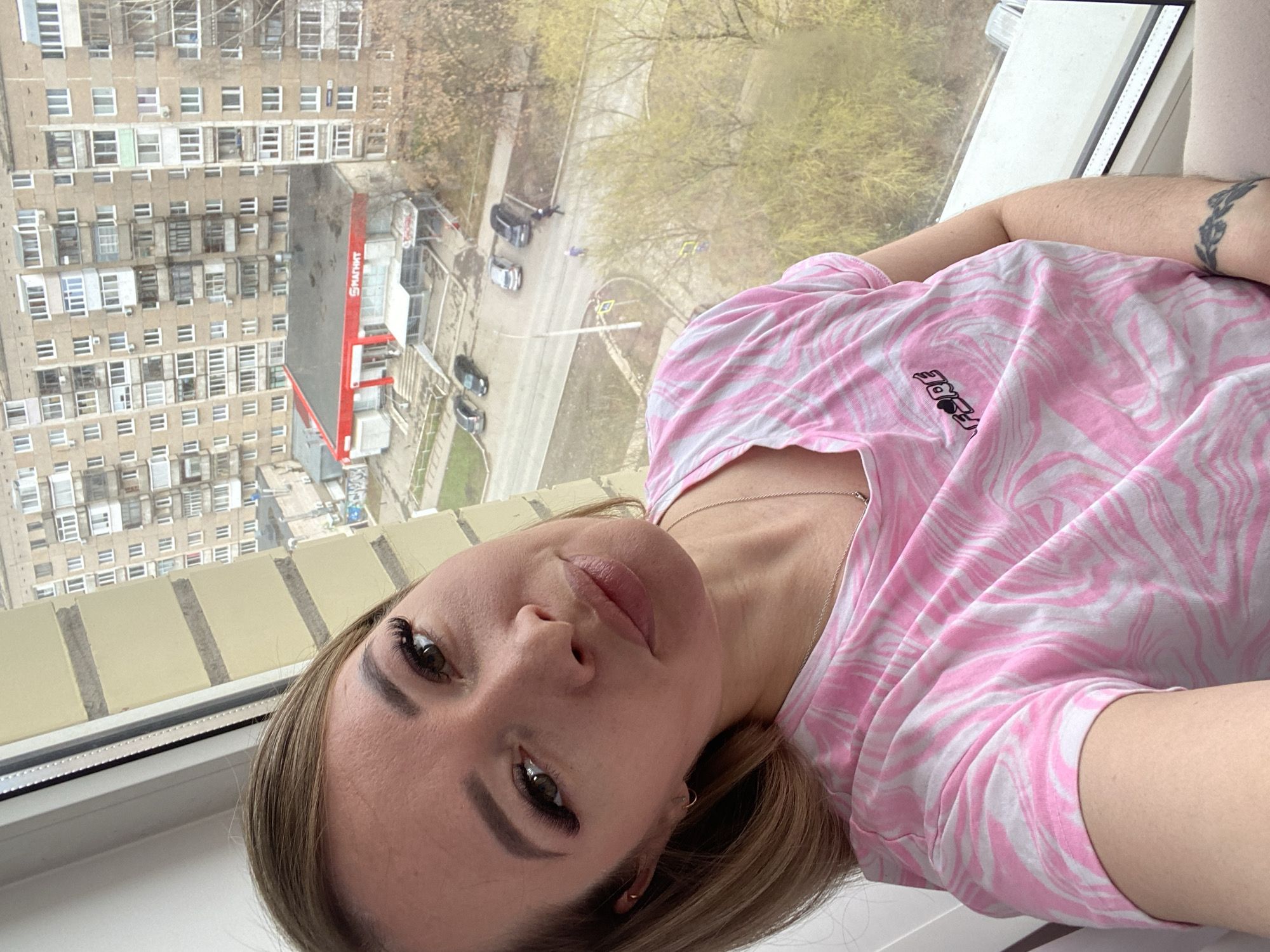 This diagonal image is a selfie of a woman taken indoors, looking out of a window. The woman, positioned in the foreground, has light brown hair, dark eyes with long eyelashes, and pink-toned lips, sporting a neutral expression. She wears a pink and white swirling patterned t-shirt, a delicate silver necklace, and a small earring. A dark band-like tattoo is visible on her upper right arm. A small, partially obscured logo on her shirt appears to read "FORE" or similar, accompanied by a small graphic.
The background, visible through the window, depicts an urban scene from a high vantage point. Dominating the middle ground is a large, multi-story residential building with numerous windows and balconies, exhibiting a style typical of Soviet-era architecture, common in Russian cities like Izhevsk. Below this building, a red-fronted store with the name "СМАРТВЭЙ" (SMARTWAY) in white Cyrillic letters is clearly visible. A street with several parked cars and trees with sparse, light-green foliage can be seen around the store and residential area.
The lighting suggests an overcast or cloudy day, with soft, diffused light and no harsh shadows. The scene evokes a sense of everyday life in a Russian city during what appears to be early spring or late autumn. The woman is likely reclining, taking the photo from an elevated position within a building.