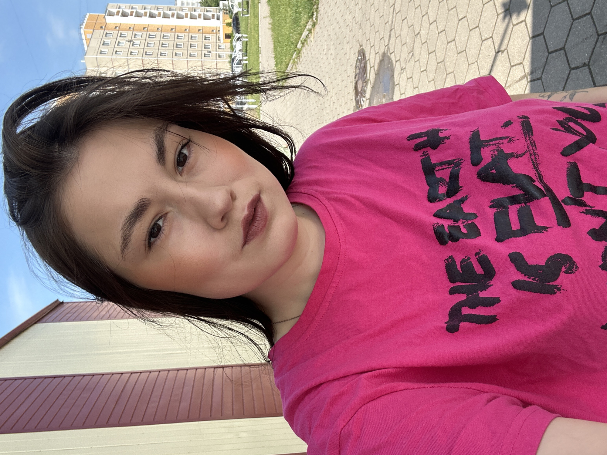 A young woman with dark hair, wearing a bright pink t-shirt with black lettering that says "THE EARTH IS FLAT", stands outdoors in front of a building with vertical red and yellow siding. The building appears to be an industrial or utility structure. In the background, a residential building with many windows and light-colored brickwork is visible, along with some parked cars and a paved walkway with a hexagonal pattern. The sky is blue and clear, with some faint clouds. The overall setting suggests a suburban or urban environment in Chaikovsky, Russia.