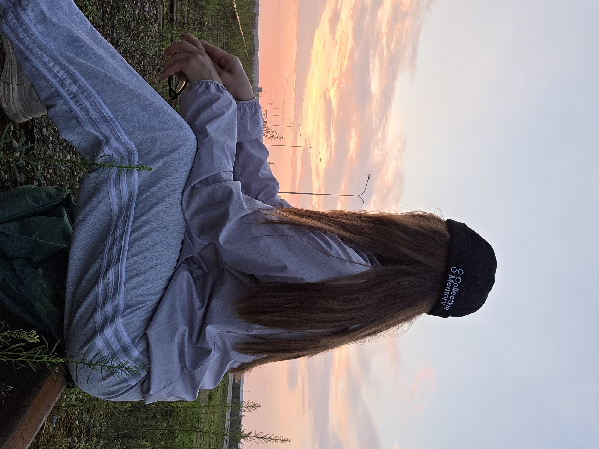 A person with long brown hair, wearing a black beanie with "Collective Memory" embroidered in white, a light grey jacket, and grey sweatpants with white stripes, sits on a railroad track. The person is facing away from the camera, looking towards a sunset with orange and pink clouds. The background features a landscape with tall grass, power lines, and streetlights. The image appears to be taken in Chaikovsky, Russia, given the railroad tracks and power lines common in industrial or semi-urban areas of the city. The overall atmosphere is one of solitude and contemplation.