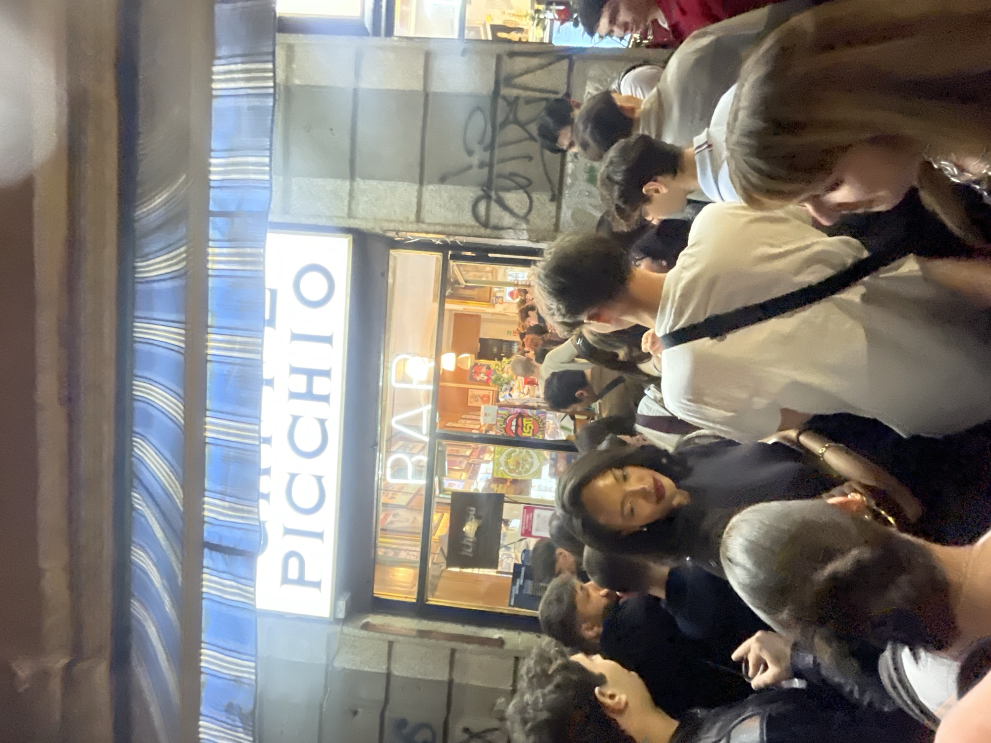 A medium shot photograph captures a crowded street scene at the "Picchio" bar in Milan, Italy. The street is filled with people, a mix of individuals standing and interacting, creating a dense gathering. The bar's signage is visible, and the interior appears busy. Graffiti covers the exterior walls. The scene is lit by the bar's lights and ambient street lighting.