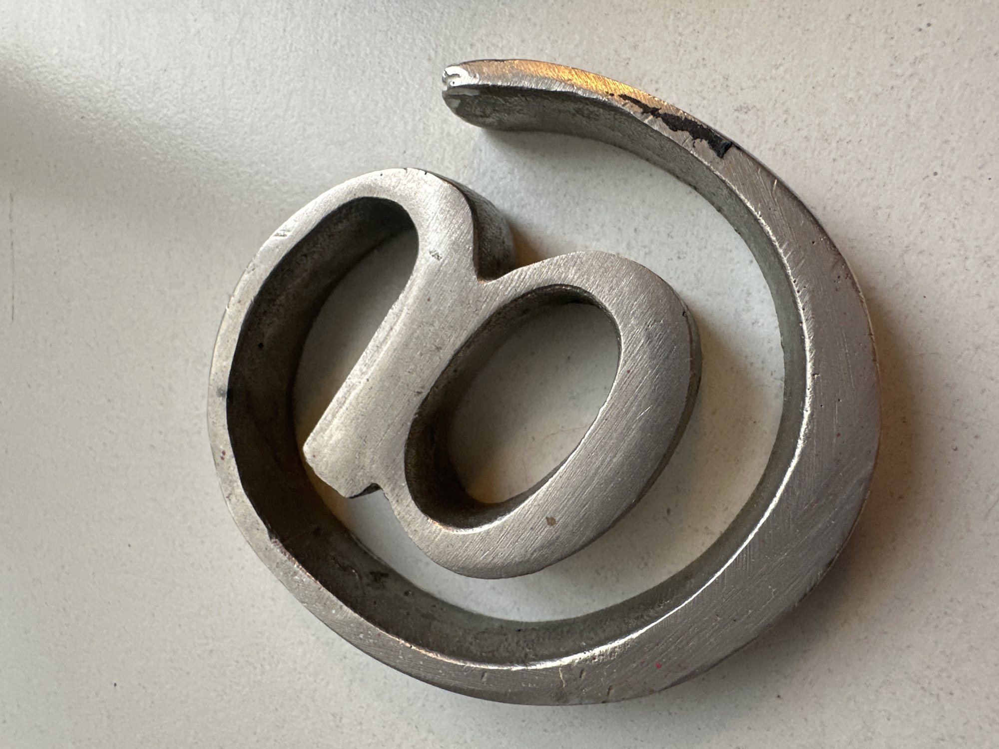 A silver-colored metal '@' sign rests on a light gray or off-white surface. The sign is three-dimensional with a somewhat rough, unfinished appearance, showing machining or tooling marks. There are a few small dark smudges or imperfections on the metal surface. The setting appears to be indoors on a neutral-colored surface. There is no indication of the location being Buenos Aires, Argentina.
