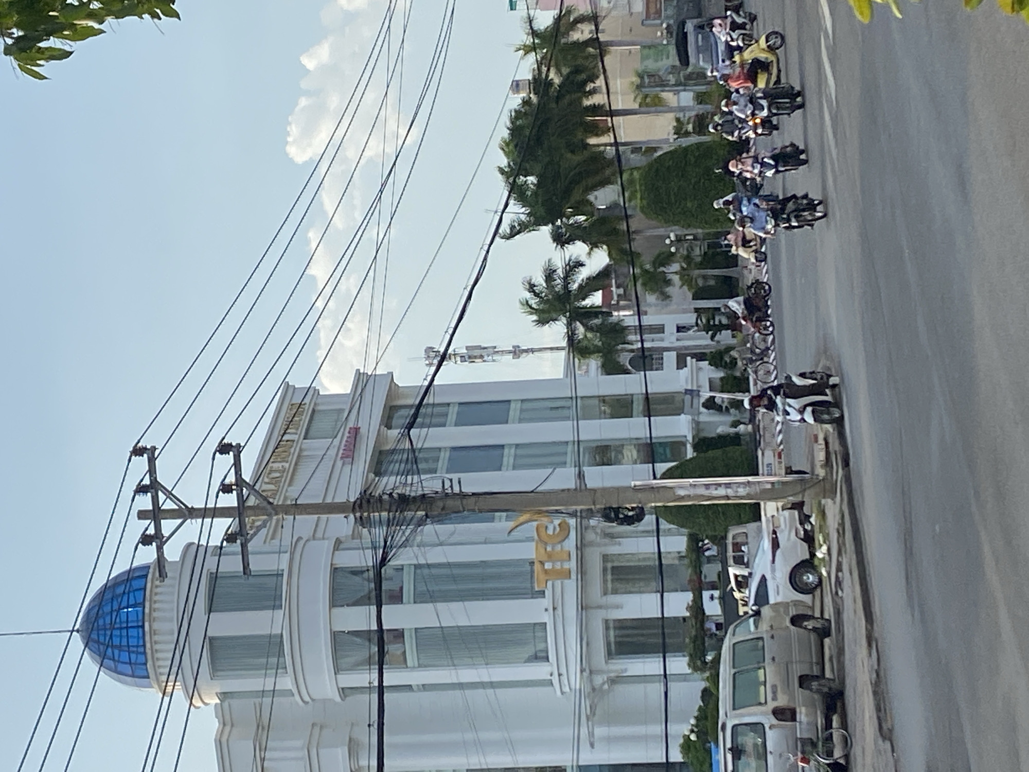 The image captures a street scene in Phan Thiết, Vietnam. A white building with a blue dome and "TFC" signage stands prominently, its architecture incorporating multiple windows and ornamental columns. Overhead power lines crisscross the sky.
A row of parked cars and motorcycles lines the right side of the street. Pedestrians and motorcyclists are visible along the roadway, suggesting an active urban environment. Palm trees punctuate the scene, and leafy foliage is present at the top left corner of the image. The sky is a clear, pale blue with a few scattered clouds.