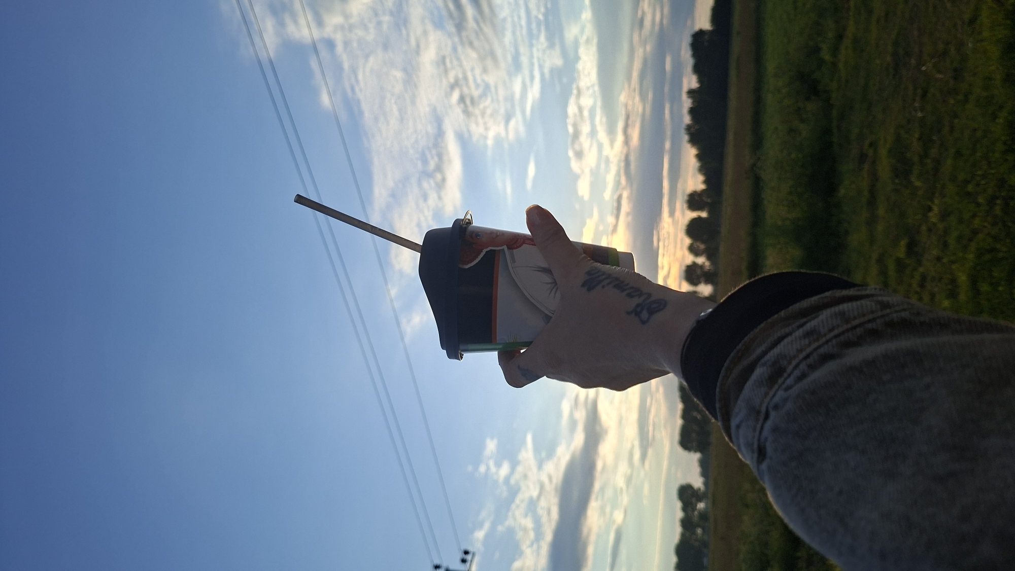 A first-person perspective shot captures a hand holding a disposable coffee cup with a straw against a backdrop of a blue sky with scattered clouds and power lines. The cup has an image of a cowboy-like character. The hand has a tattoo on the knuckles that reads "Vanille" and another abstract tattoo on the back of the hand. The person is wearing a denim jacket. In the background, a grassy field stretches towards a line of trees under the setting or rising sun, suggesting an outdoor rural setting. The scene appears to be in the Zabalotsky rural council of Belarus.