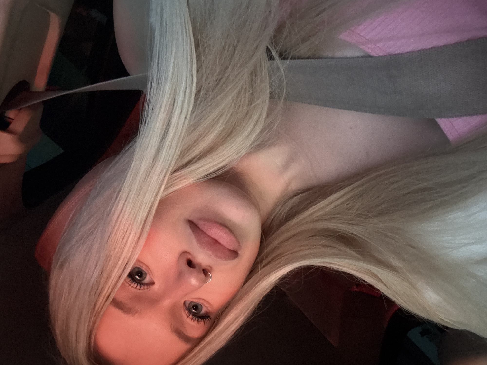 A young woman with long blonde hair, wearing a pink top and a grey seatbelt, is captured in a close-up, candid selfie. Her face is illuminated by warm, orange light, casting a rosy hue on her skin. She has a subtle smile, her eyes are looking towards the camera, and she is wearing a small silver hoop in her nose. The background is dark, with hints of red and blue lights, suggesting she might be in a vehicle at night. The image quality is grainy, typical of a low-light smartphone photo. No specific location in Saint Petersburg can be identified from the visual information alone.