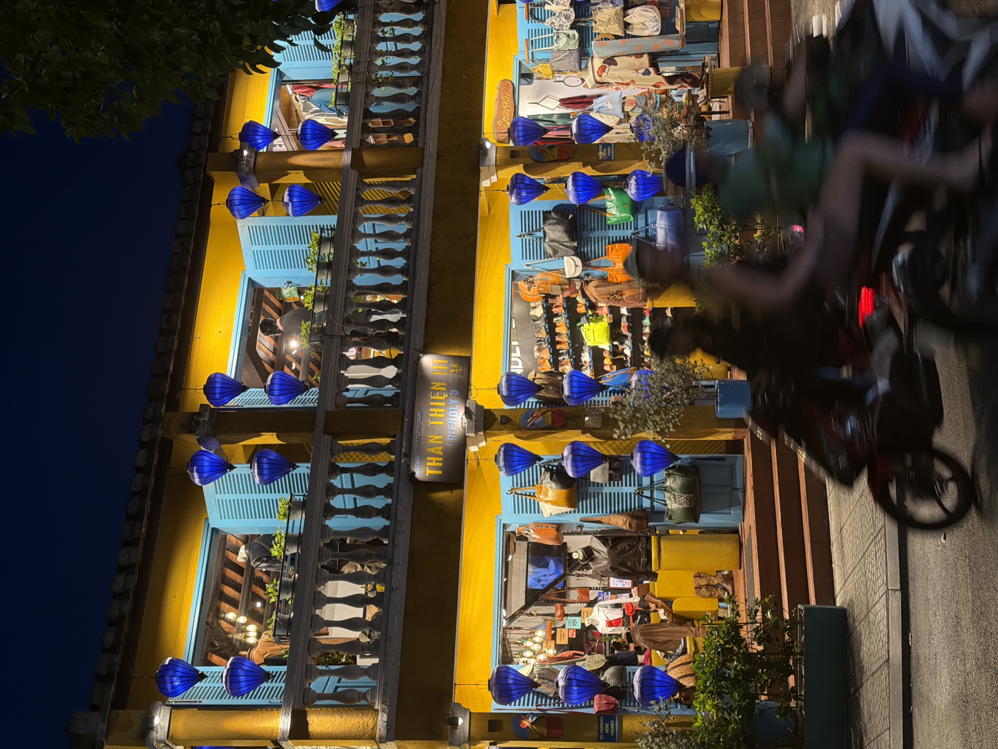 In Hoi An, Vietnam, at night, a yellow building with blue shutters and balconies is illuminated. Blue lanterns hang from the balconies and in front of the shop windows. The ground floor of the building houses a shop selling bags, shoes, and clothing. A blurred image of two people on a motorcycle passes in front of the shop. The street is wet, suggesting recent rain.