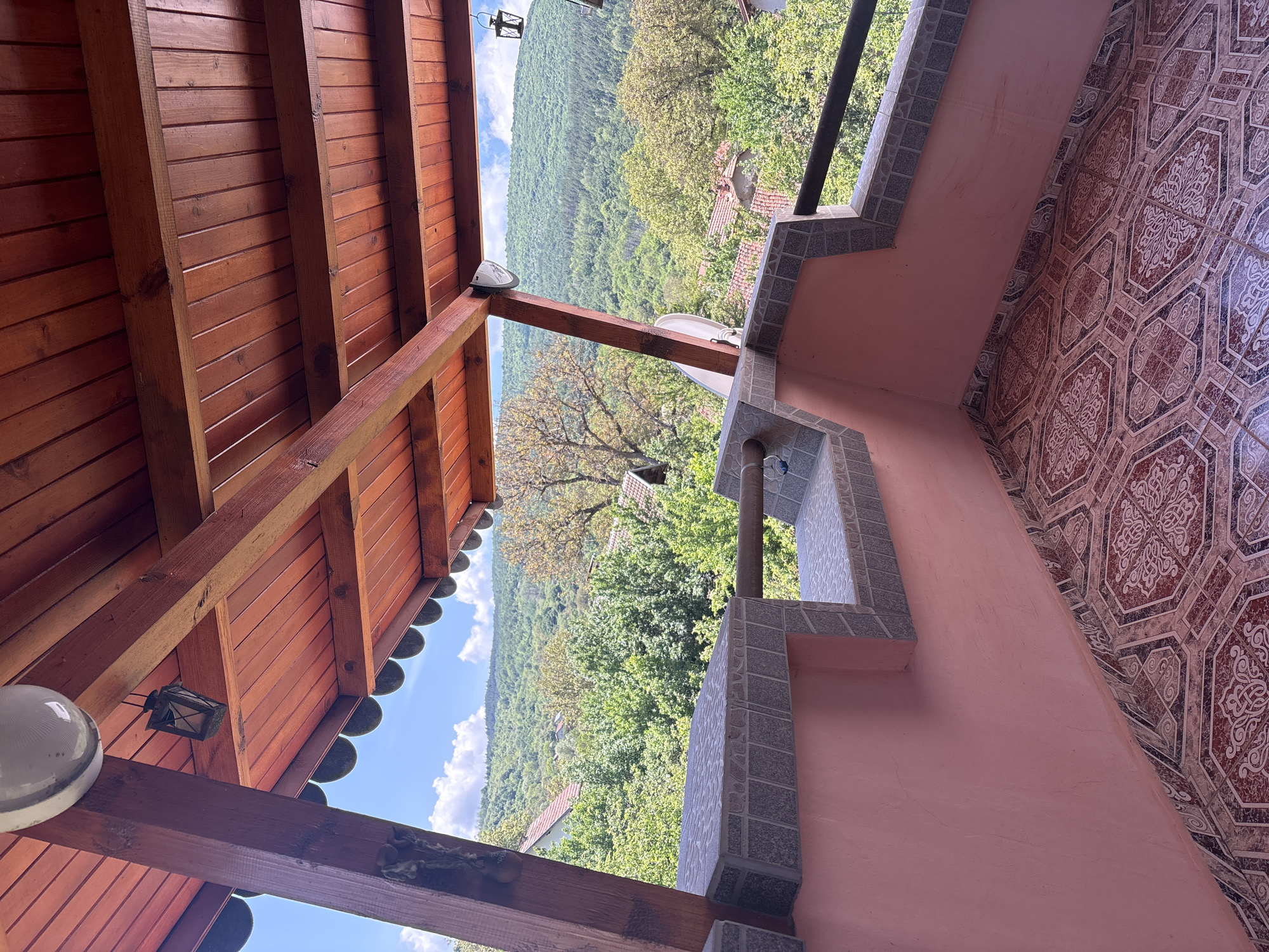 The image captures a panoramic view from a covered outdoor area, likely a balcony or porch, overlooking a vast forested valley in Svetlia, Bulgaria. The scene is illuminated by bright daylight under a clear blue sky adorned with scattered white cumulus clouds.
On the left side of the frame, a rustic wooden ceiling is prominently featured, constructed from reddish-brown planks supported by sturdy wooden beams. Two distinct light fixtures are attached to the ceiling: a modern, spherical white globe light and an older, black-framed lantern.
The right side of the image reveals a salmon-pink wall paired with an ornate tiled surface, possibly a floor or another wall section, showcasing intricate geometric and curvilinear patterns in shades of red, brown, and cream. A decorative balustrade or low wall, built from greyish-white textured tiles, forms a barrier, from which a black metal railing extends. A small satellite dish is mounted below the railing, and a water tap with associated piping is visible on the tiled structure.
The middle ground and background are dominated by a sprawling, dense green forest that covers numerous hills and valleys, stretching towards the horizon. Nestled among the trees, several houses with characteristic red or orange tiled roofs are visible, indicating a rural settlement within the natural landscape.
The overall impression is one of serene natural beauty and a peaceful rural setting. No people are visible in the image, and there is no discernible text.