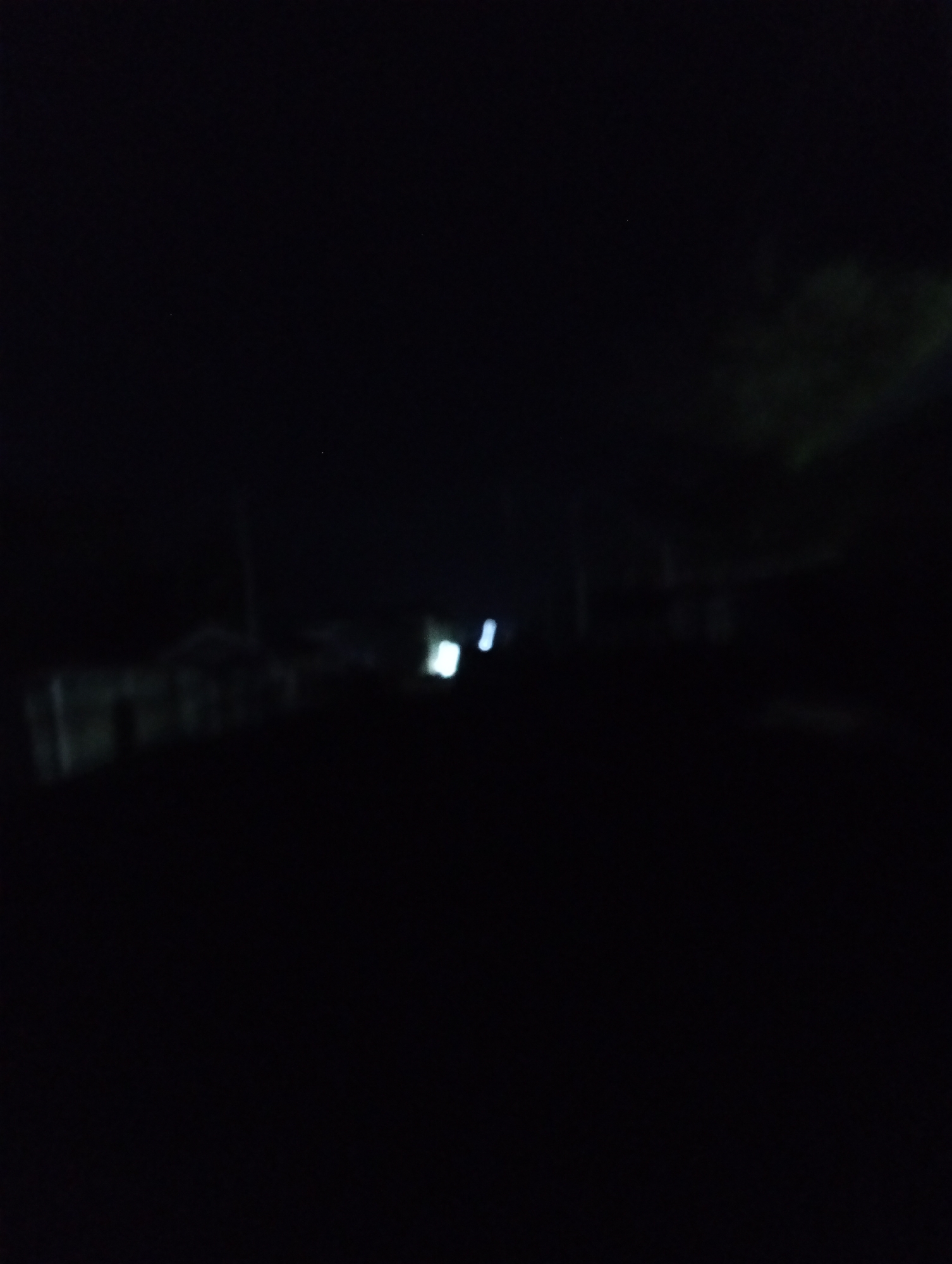 A dark street scene at night in Adankolo, Nigeria. Barely visible are buildings on the left and right. There are a couple of faint lights further down the street. Foliage is on the right side of the image. The foreground is dark.