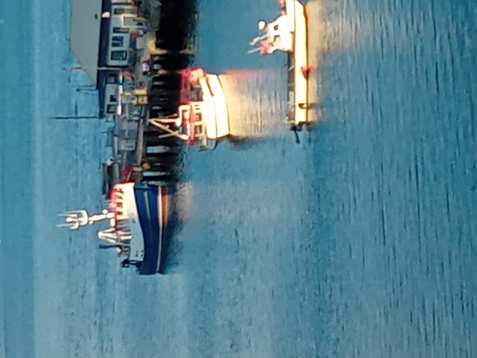 The image shows a harbor with several boats docked and at anchor. The water is calm and reflects the blue sky. In the foreground, there is a small, white boat with a red fender. Behind it, a larger, white fishing boat with red trim is anchored. Further back, a blue and white fishing boat is docked at a pier. A building with a light-colored roof and dark windows is visible on the pier. The overall scene depicts a typical day in a fishing harbor.