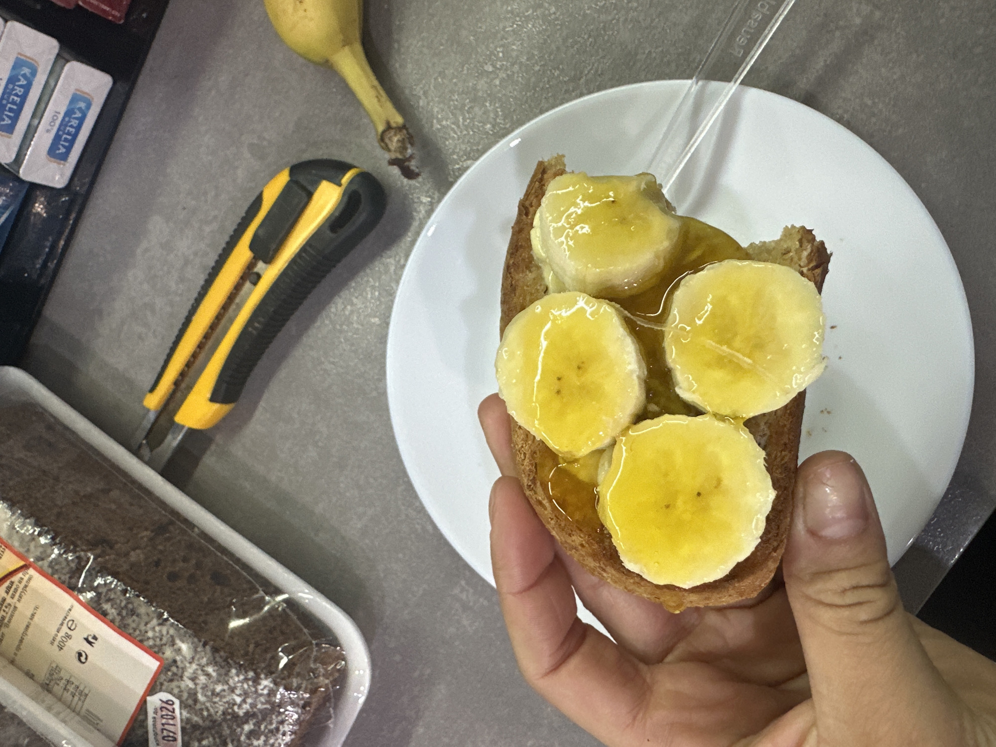 A person's hand is holding a slice of bread topped with sliced bananas and drizzled with honey. The bread is on a white plate, which is placed on a gray countertop. In the background, to the left, is a loaf of dark bread in plastic packaging, with a sticker labeled "071026". Next to the bread packaging is a yellow and black utility knife. To the right of the knife is a partially visible banana. Further in the background, on a shelf, are several small packages with blue and white labels that appear to be butter or cheese. The scene is consistent with a home kitchen environment, and based on the provided context, this location is Malevo, Bulgaria.